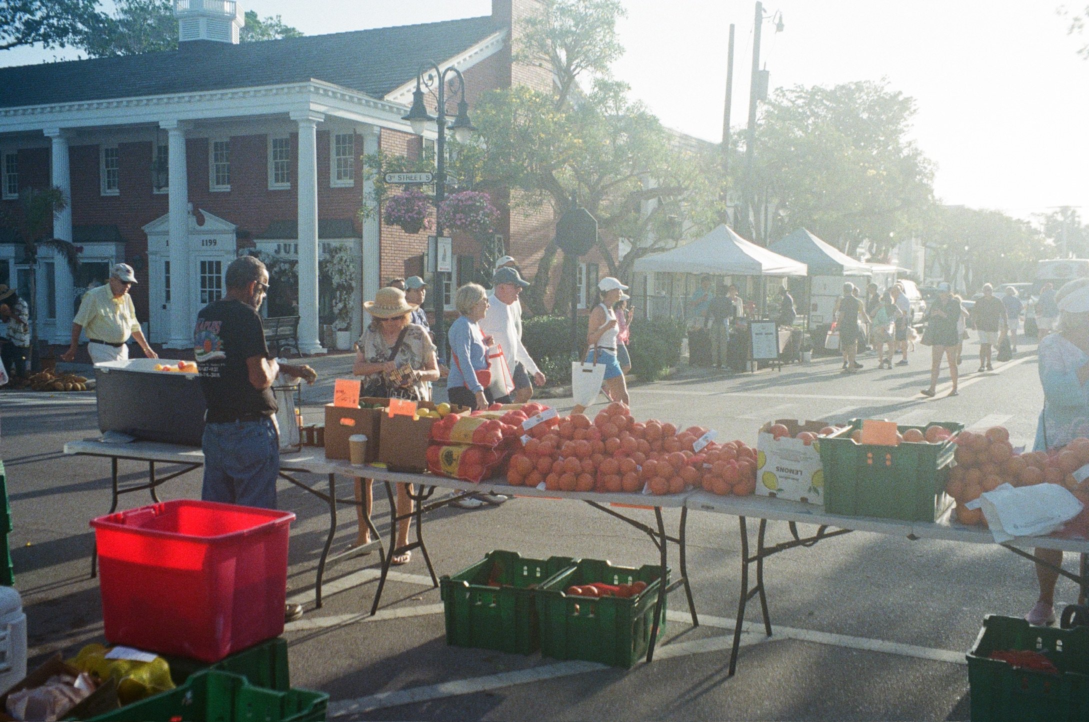 People shopping at an outdoor farmer's market with tables of fresh fruits and vegetables on a street in a small town, with buildings, tents, and trees in the background.