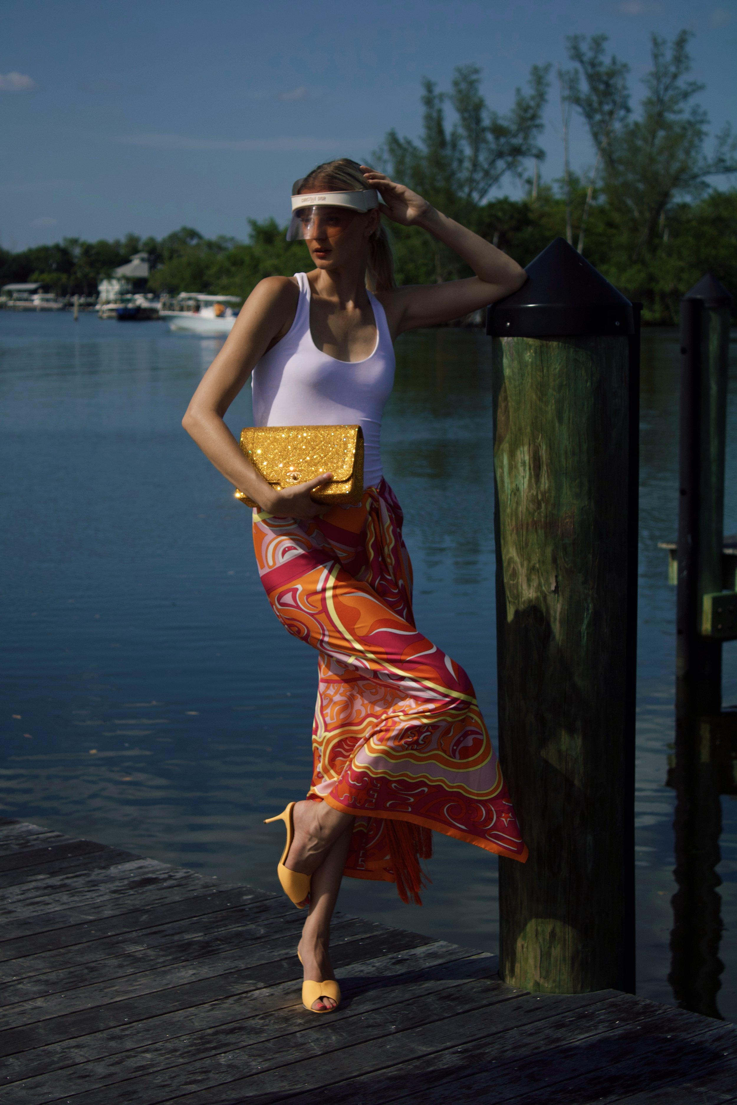 a model stands on a pier wearing a dior visor, pucci orange skirt, and holding a gold embellished chanel bag