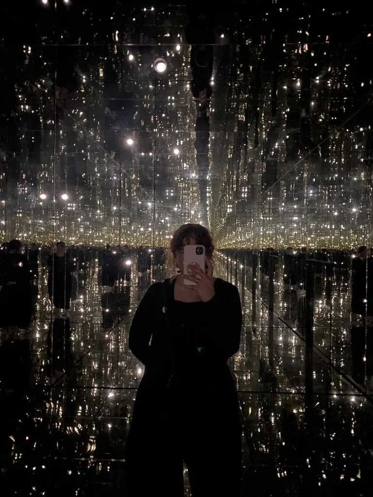 Person taking a mirror selfie in a reflective, mirrored room with numerous small lights creating a starry effect.