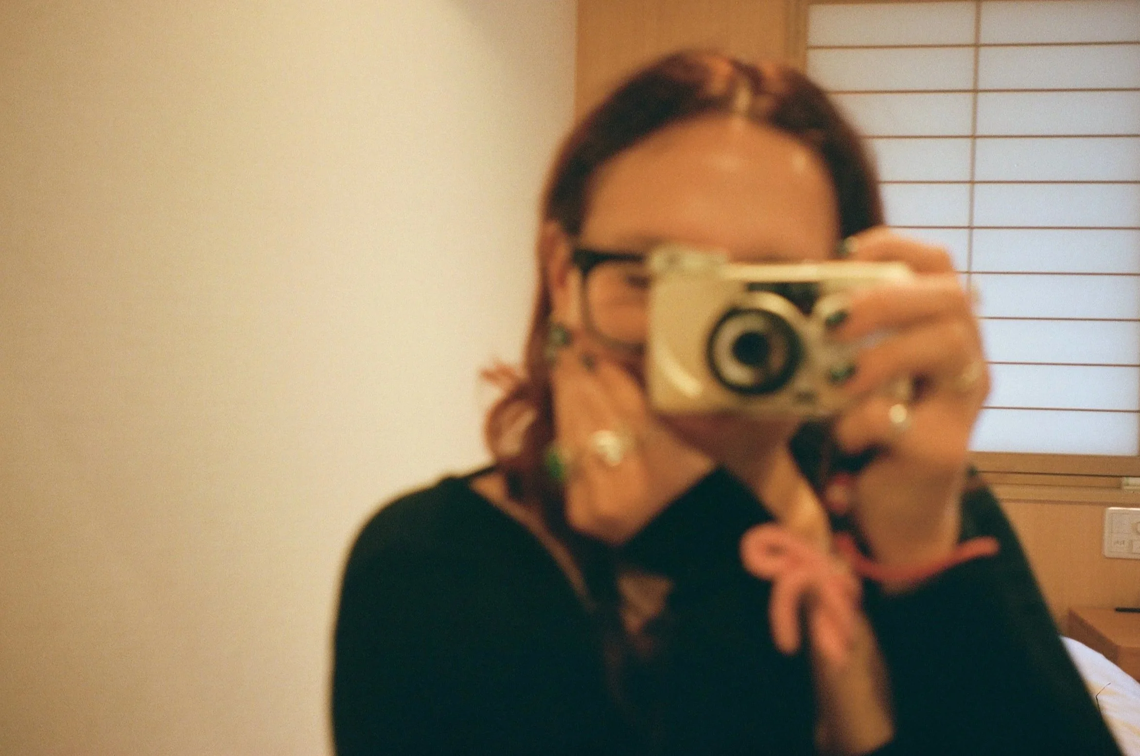 A person taking a mirror selfie with a digital camera. The person has dark hair, glasses, and is wearing a black shirt. The background shows a plain wall and a wooden panel with horizontal lines.