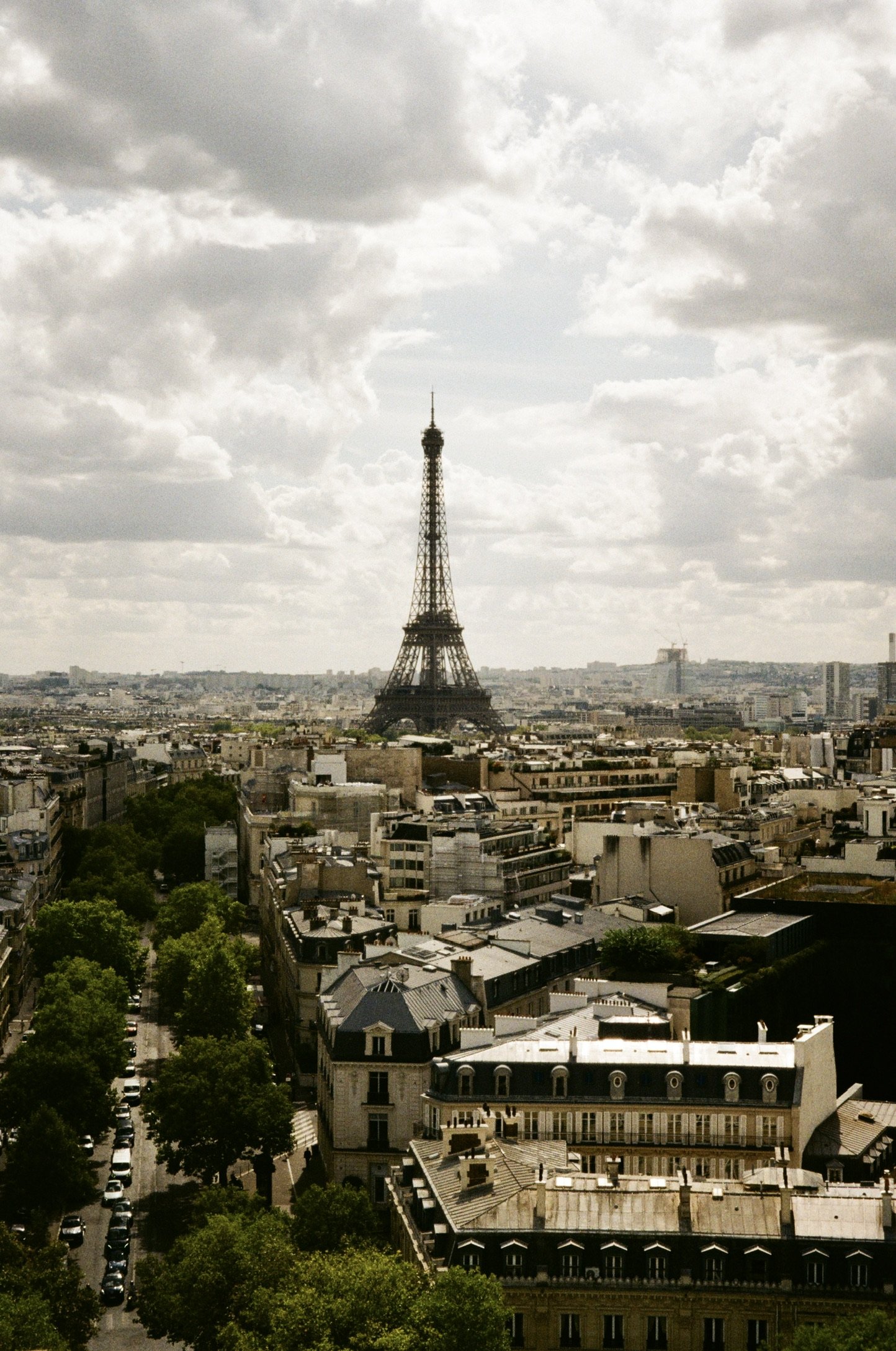 A cityscape view of Paris, France with the Eiffel Tower in the center, surrounded by buildings and trees, under a partly cloudy sky.