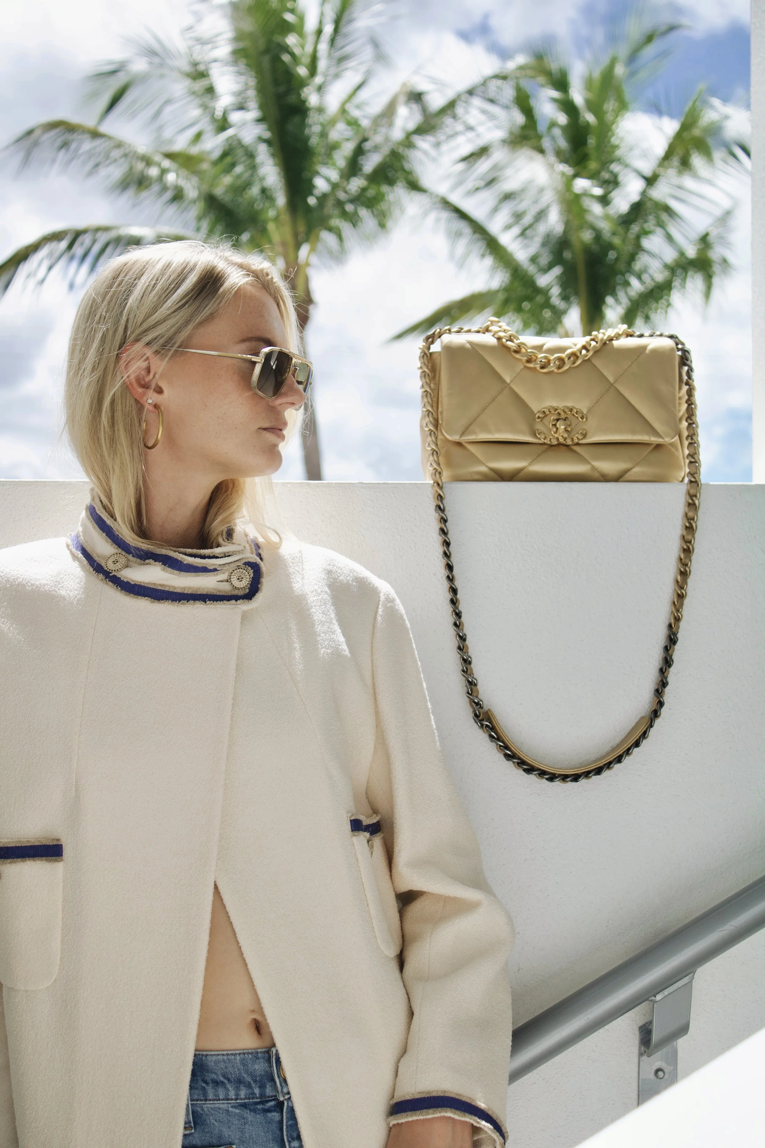 a model wears sunglasses and a long chanel coat with a gold chanel 19 flap bag next to her and palm trees in the background