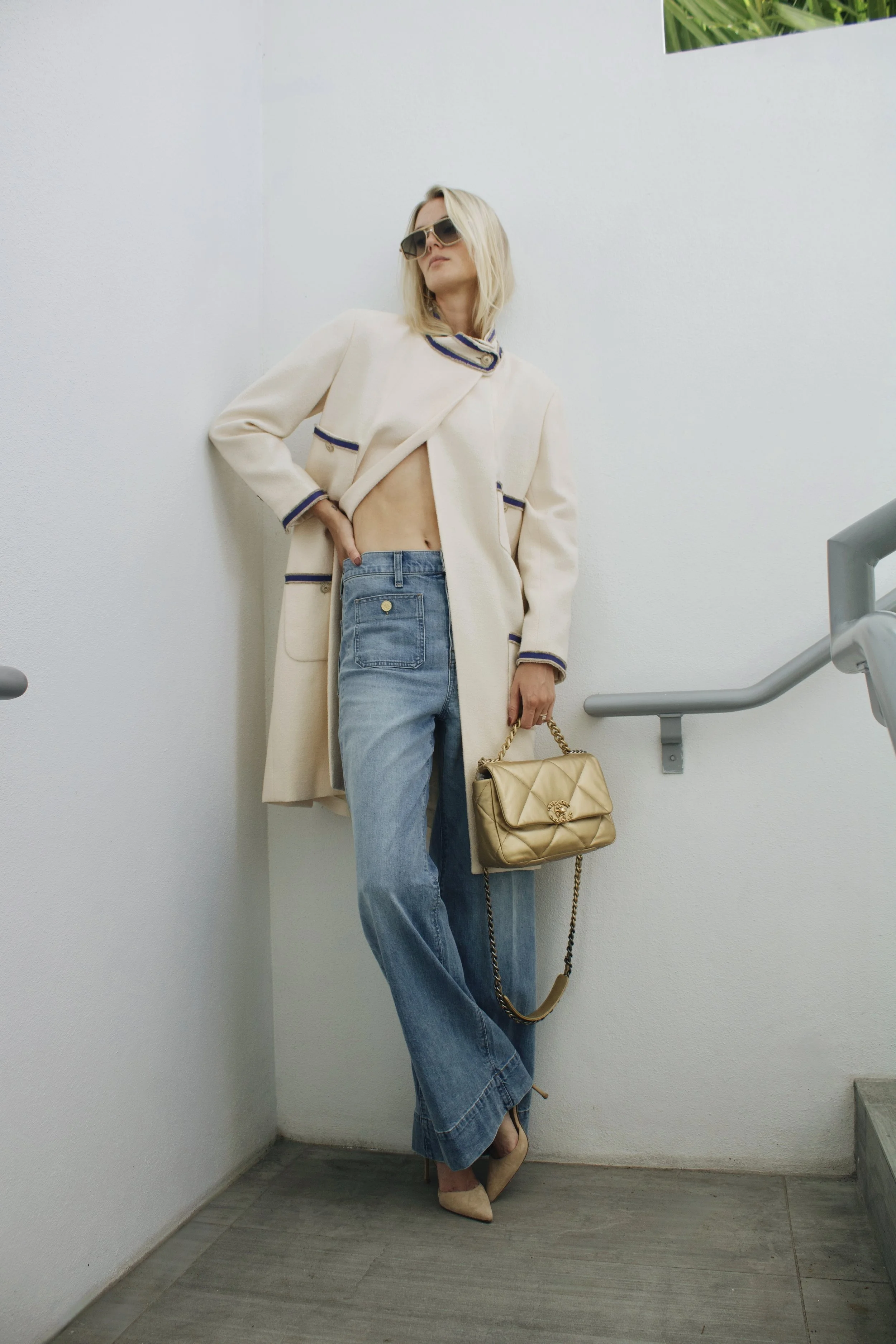 a model stands in the hallway wearng a chanel long coat, sunglasses, and jeans
