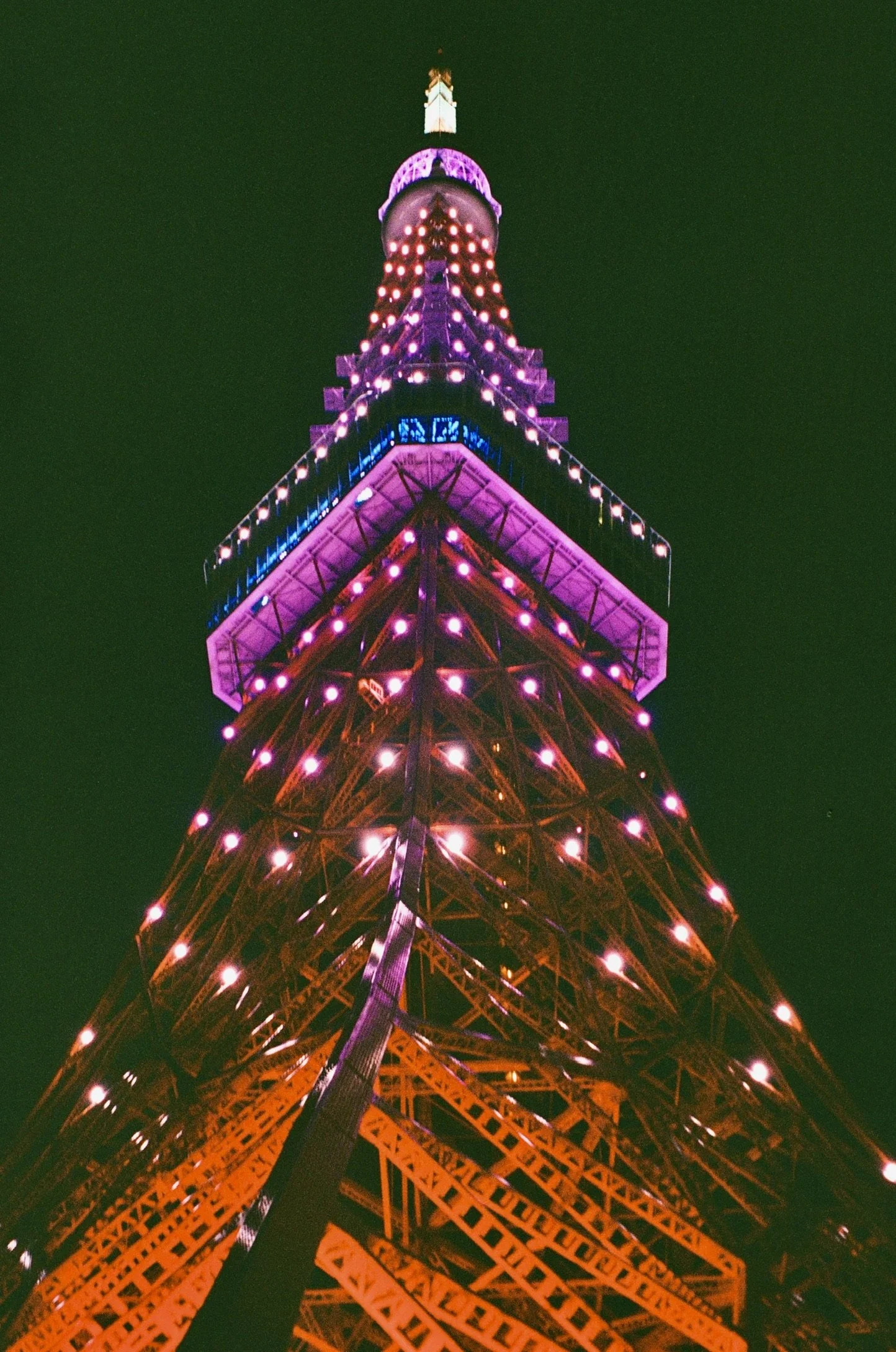 Looking up at Tokyo Tower illuminated with pink, purple, and orange lights at night against a dark sky.