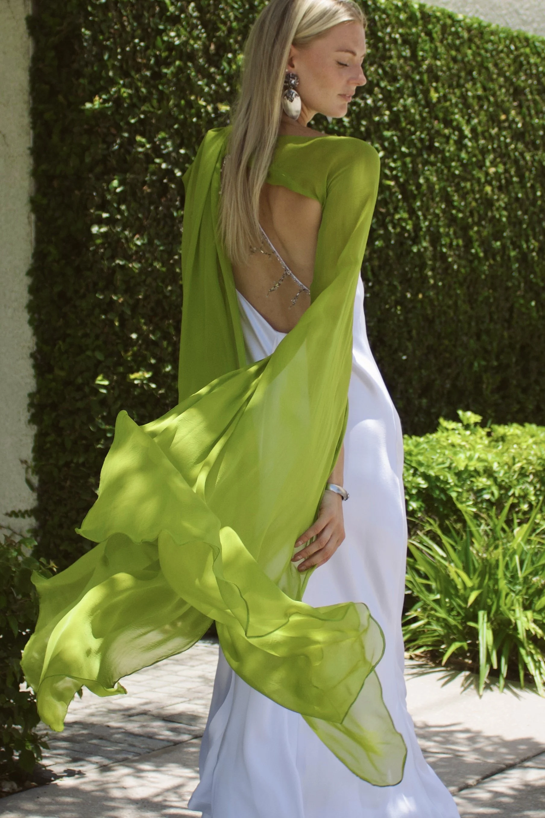 a model wearing a white dress and green chiffon scarf walks by with greenery behind her