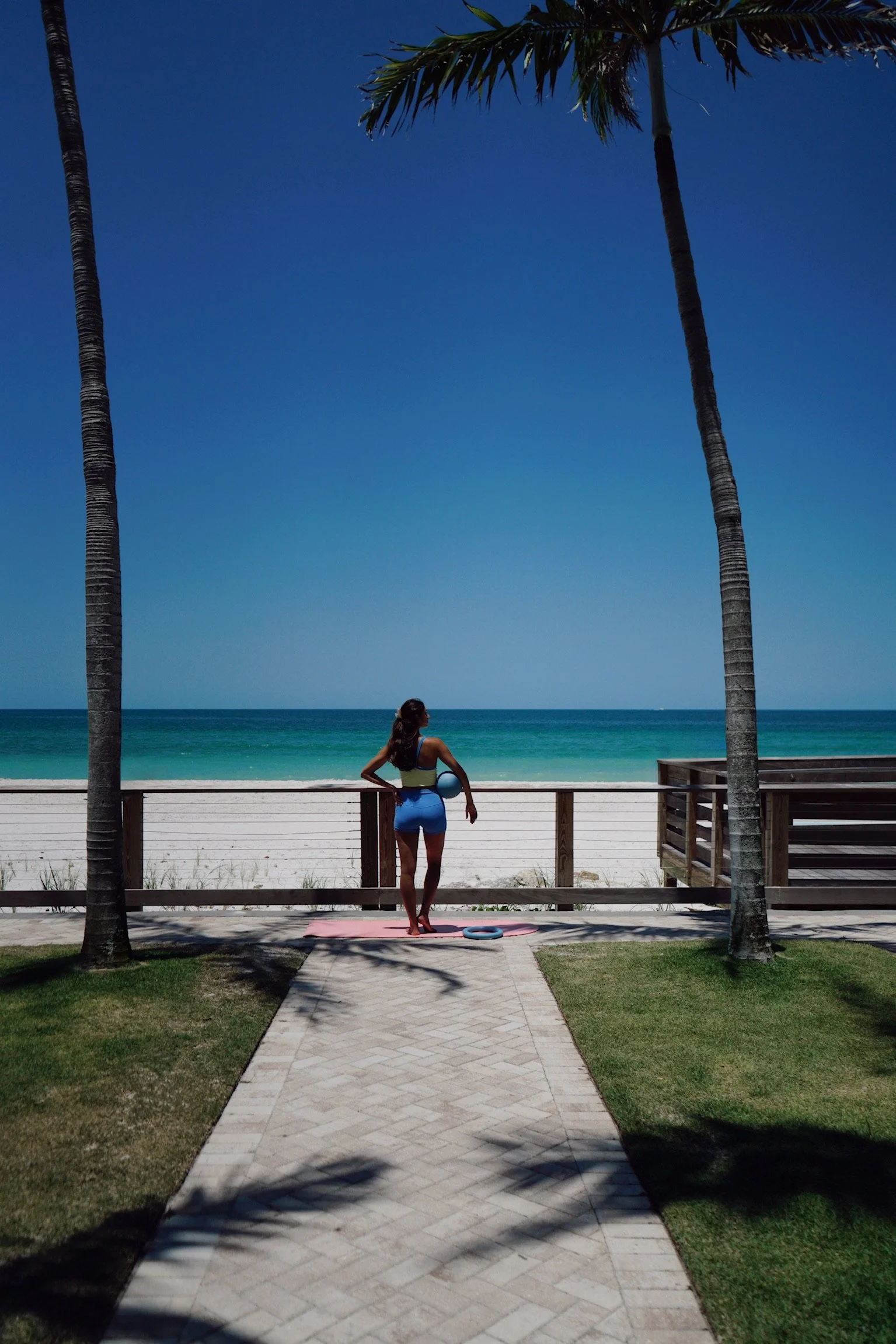 A woman practicing yoga by the beach, standing on a pink yoga mat, holding a blue ball, with two tall palm trees, a fence, and the ocean in the background on a sunny day.