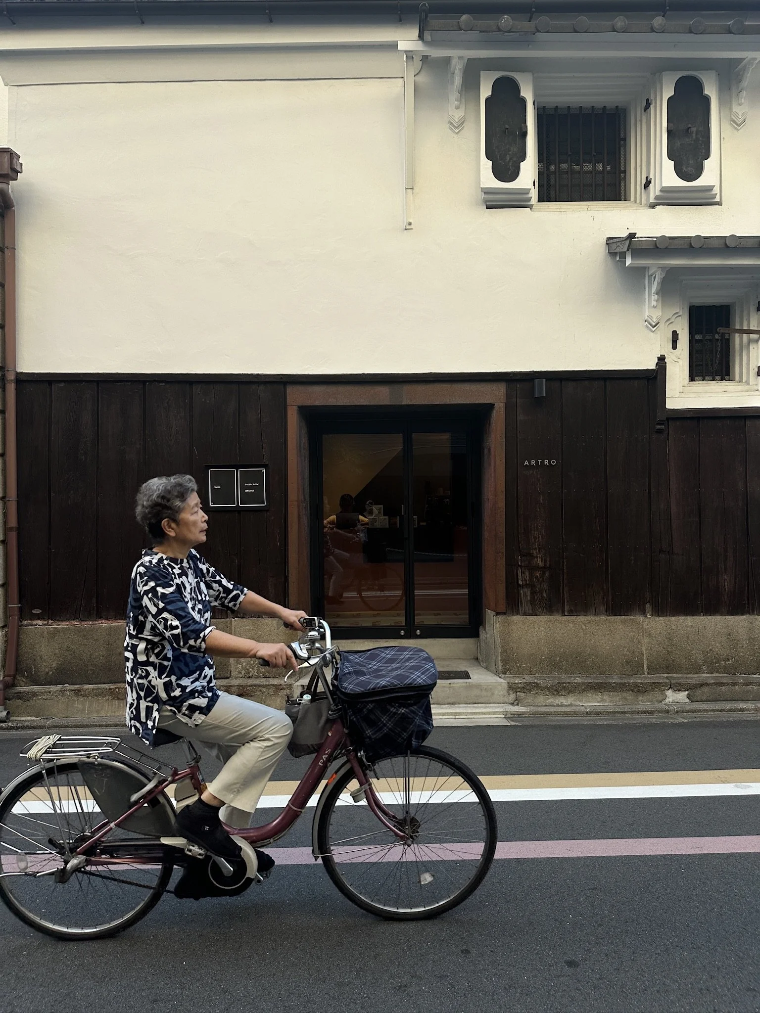 A woman with short gray hair riding a maroon bicycle on a street. She is wearing a black and white patterned shirt, beige pants, and black shoes. The street has a black and white bike lane stripe. Behind her, there is a building with a dark wooden lower section, a white upper section with windows, and an entrance door with a glass panel. The woman appears to be exiting or approaching a storefront named "ARTRO."