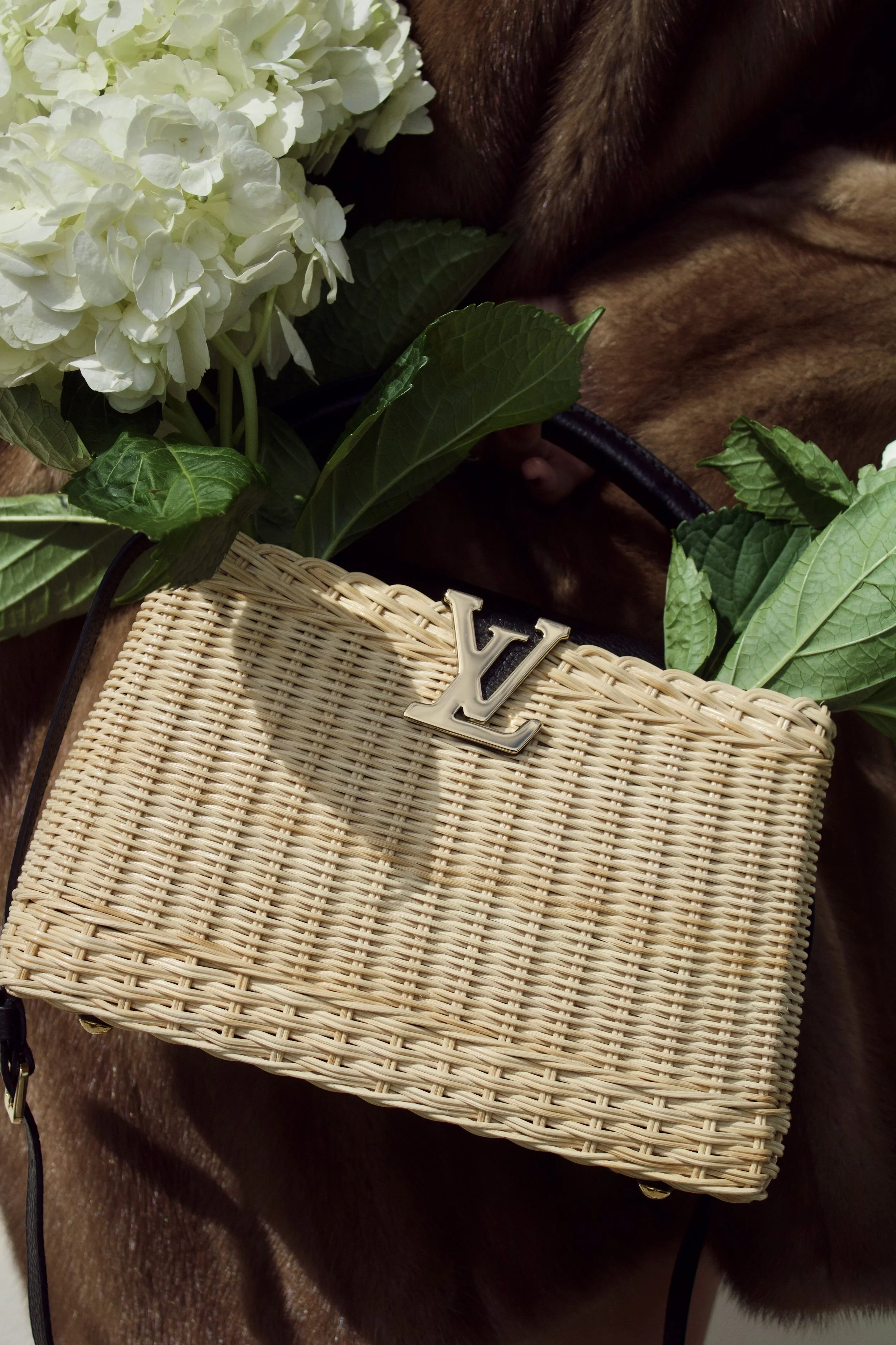 a model wearing a vintage fur coat holds a louis vuitton wicker capucines bag filled with hydrangeas