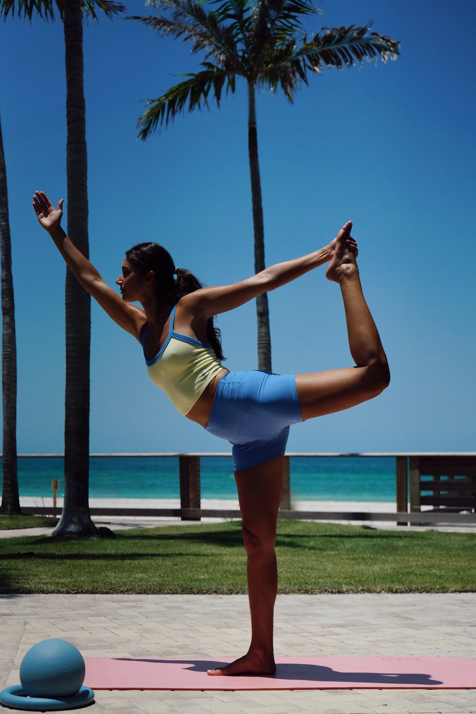 A woman practicing yoga outdoors near the beach with palm trees in the background, standing on one leg while holding her other foot behind her, with a pink yoga mat and a blue exercise ball nearby.