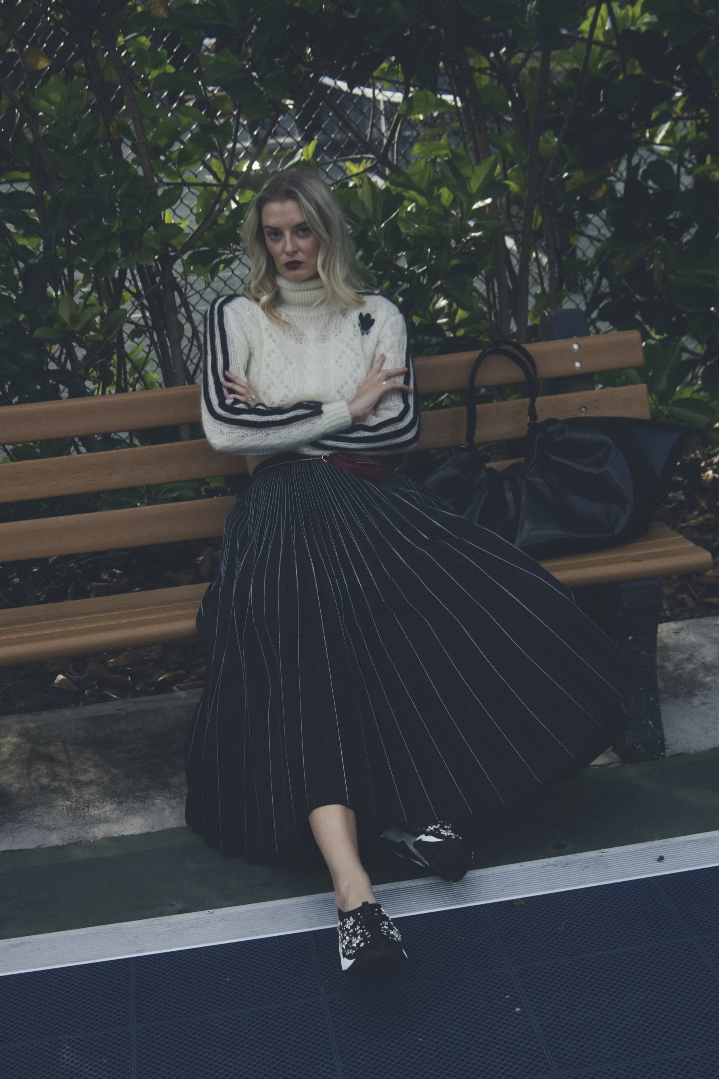 a model sits on a bench wearing a gucci sweater, black pleated skirt, fendi mini bag, and dior sneakers