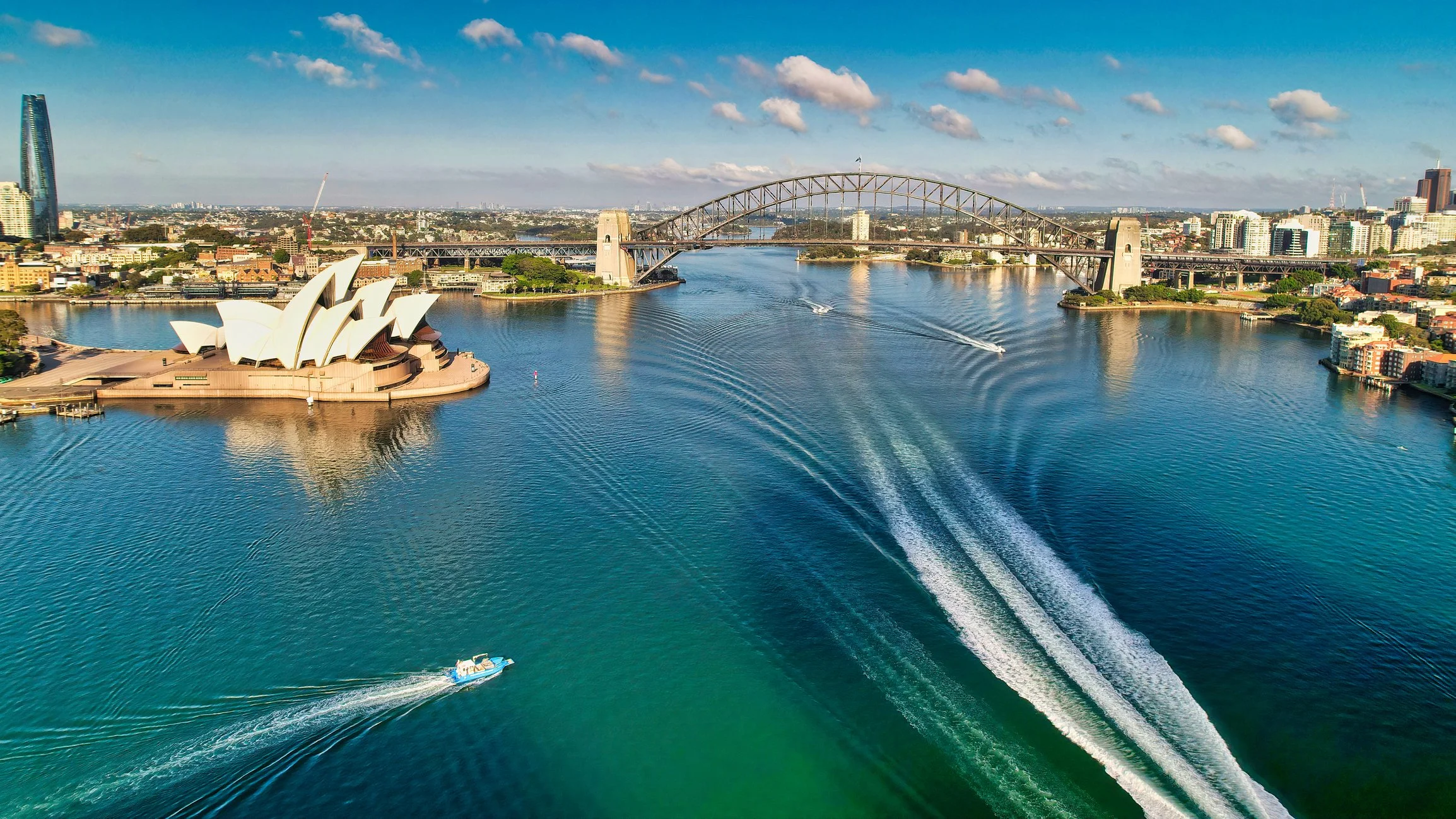 Aerial view of Sydney Harbour showing the Sydney Opera House, the Harbour Bridge, and boats on the water with a city skyline in the background under a partly cloudy sky.