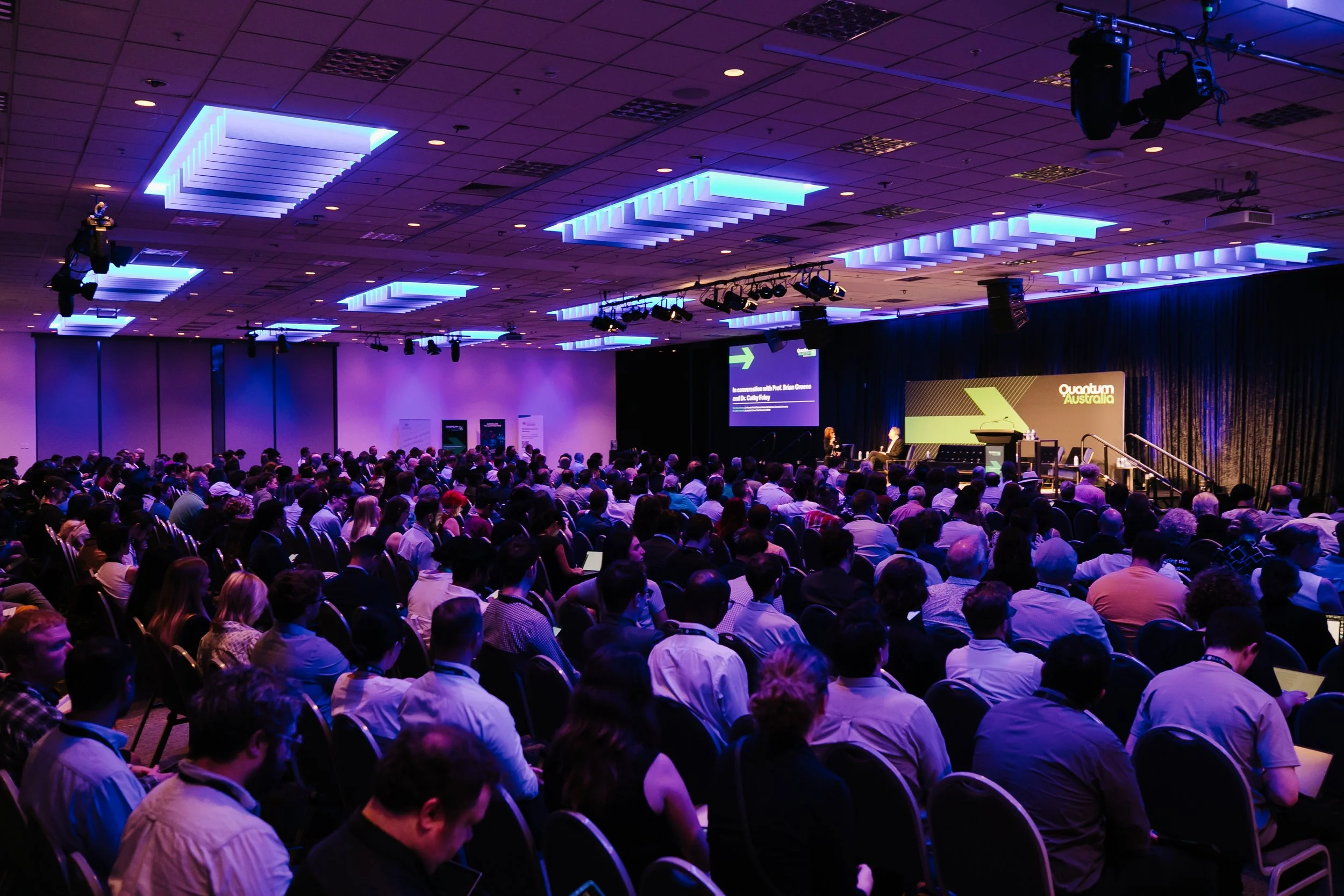 Large conference room filled with an audience of people watching a stage presentation at the Quantum Australia conference, with blue lighting on the ceiling and a projector screen on the stage.