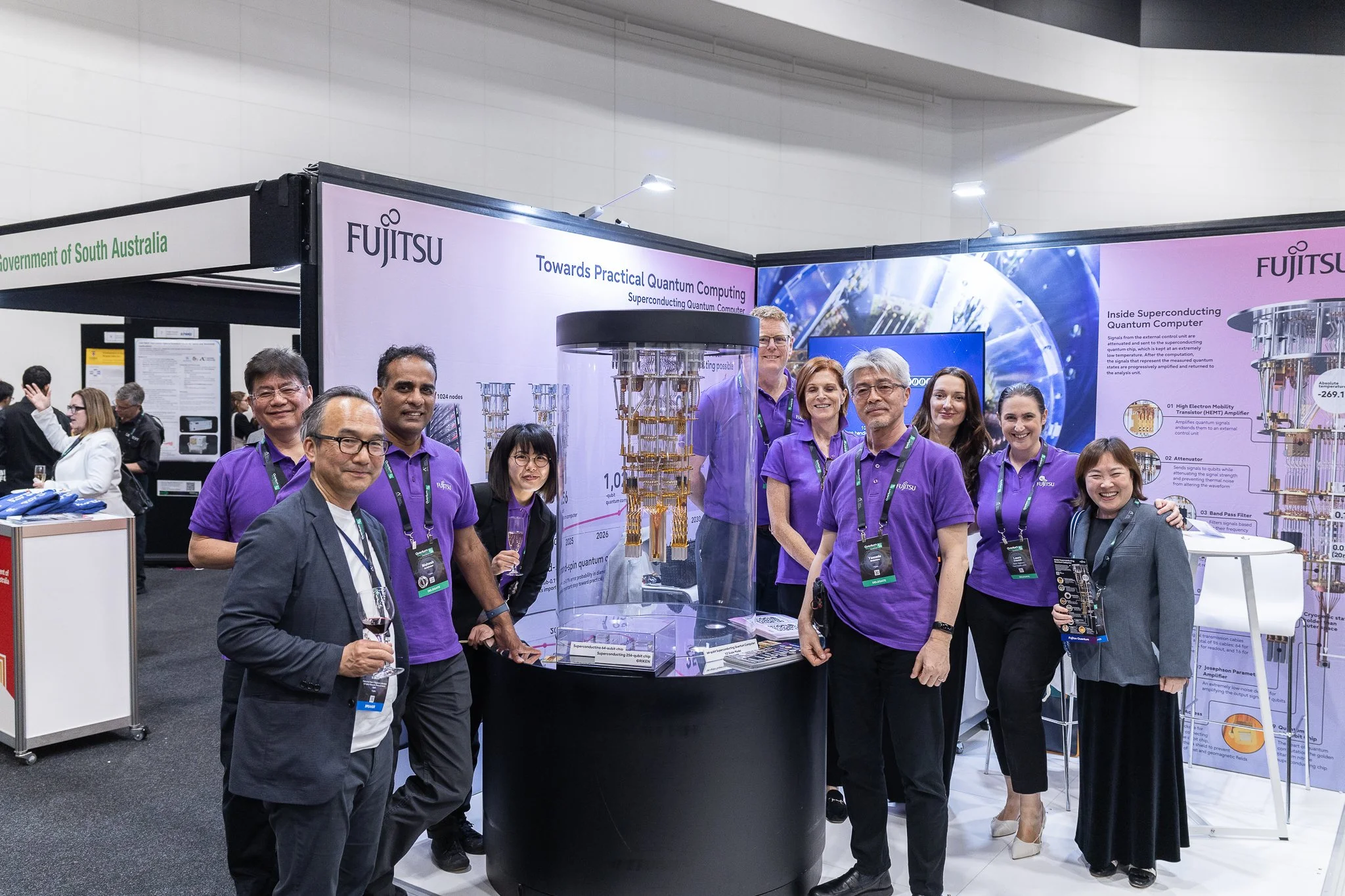 Group of people at a trade show booth for Fujitsu, showcasing quantum computing technology, with posters and displays about superconducting quantum computer