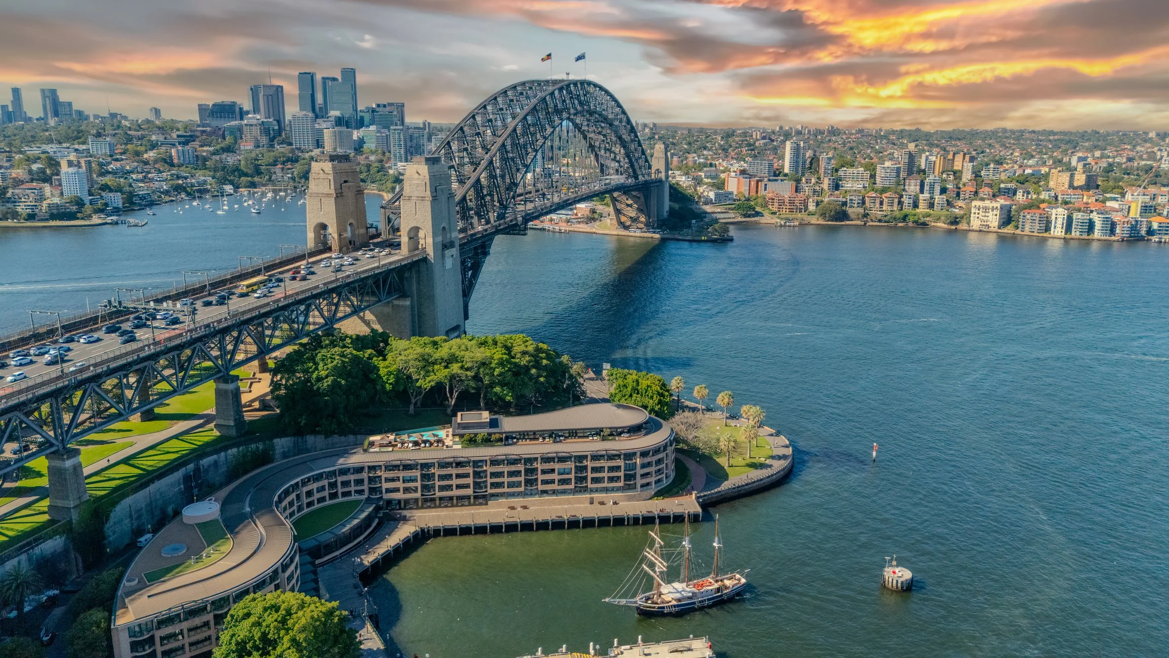 A view of the Sydney Harbour Bridge spanning across the harbor with a city skyline in the background during sunset, boats in the water, and a large curved building with a swimming pool and trees in the foreground.