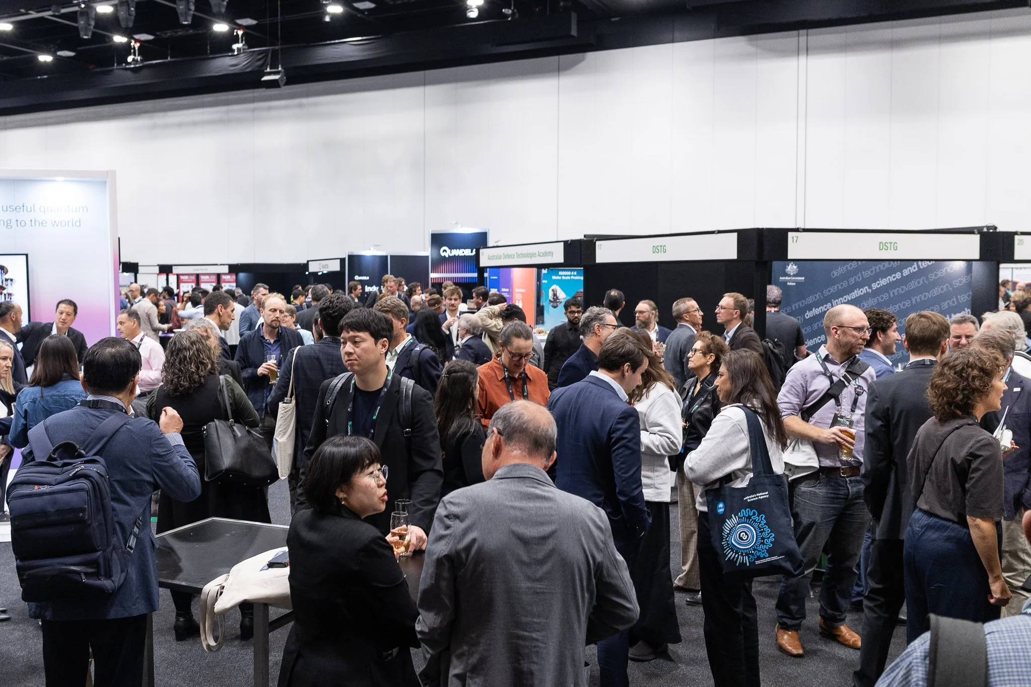 Crowd of people attending a Quantum Australia Conference trade show in a large indoor venue. Many individuals are standing, talking, or walking near exhibition booths with signs and displays.