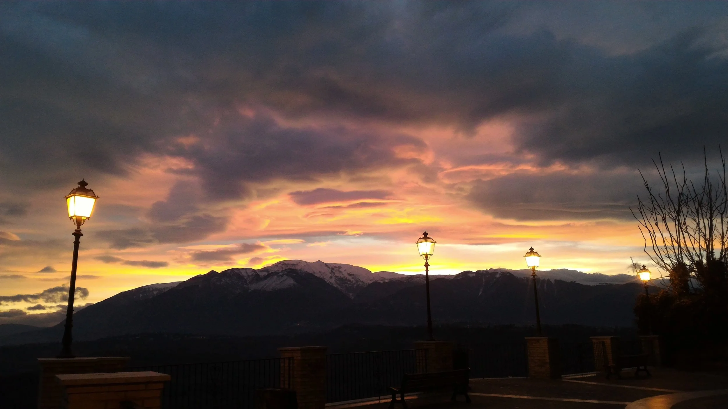 Tramonto dal belvedere di Orsogna, con nuvole scure nel cielo, montagne con neve in lontananza, lampioni accesi.