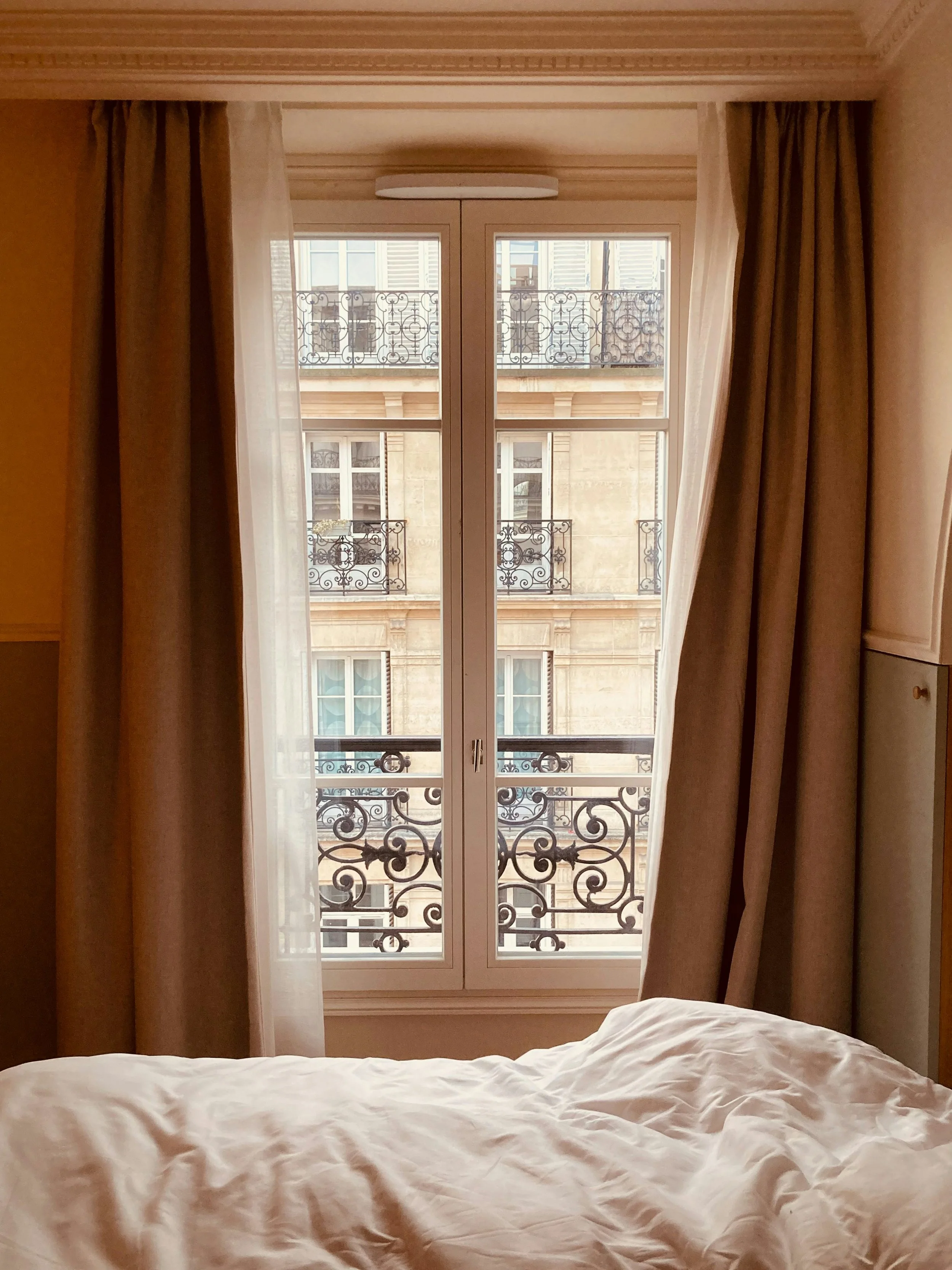 A view of a balcony through a window, with ornate black railings and a neighboring building with balconies and windows, partially covered by white and beige curtains, from a bed with a white blanket.