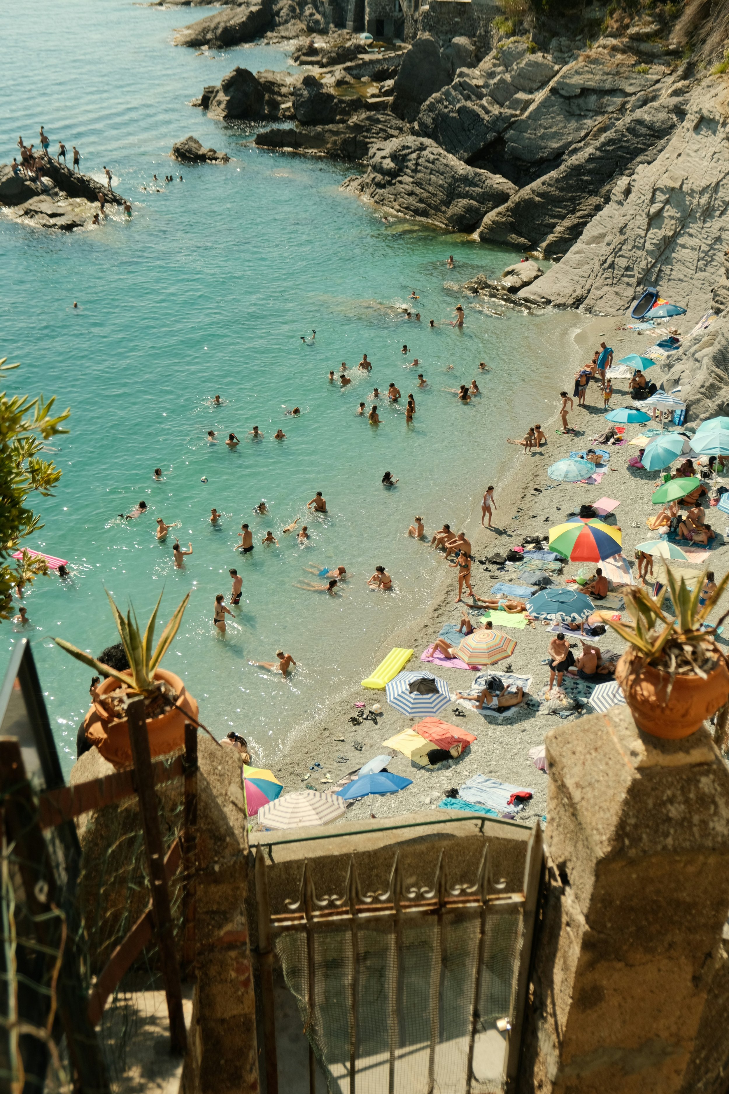 A crowded beach with numerous people swimming and relaxing under umbrellas, rocky cliffs surrounding the shoreline, and a staircase leading down to the water.
