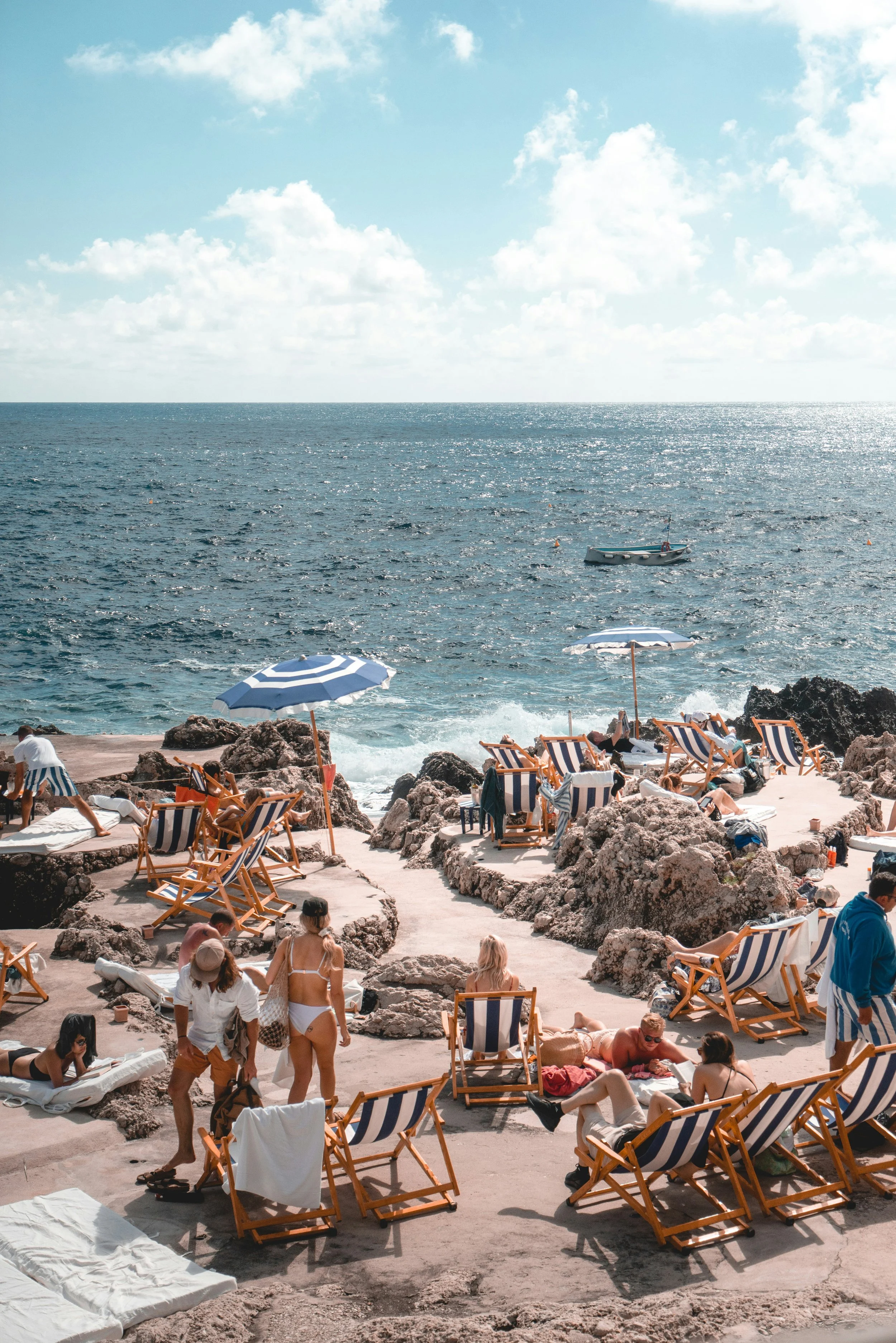 A sunny beach scene with people relaxing on lounge chairs and walking along a rocky shoreline. The ocean is in the background with small boats and umbrellas are scattered around the sandy and rocky area.