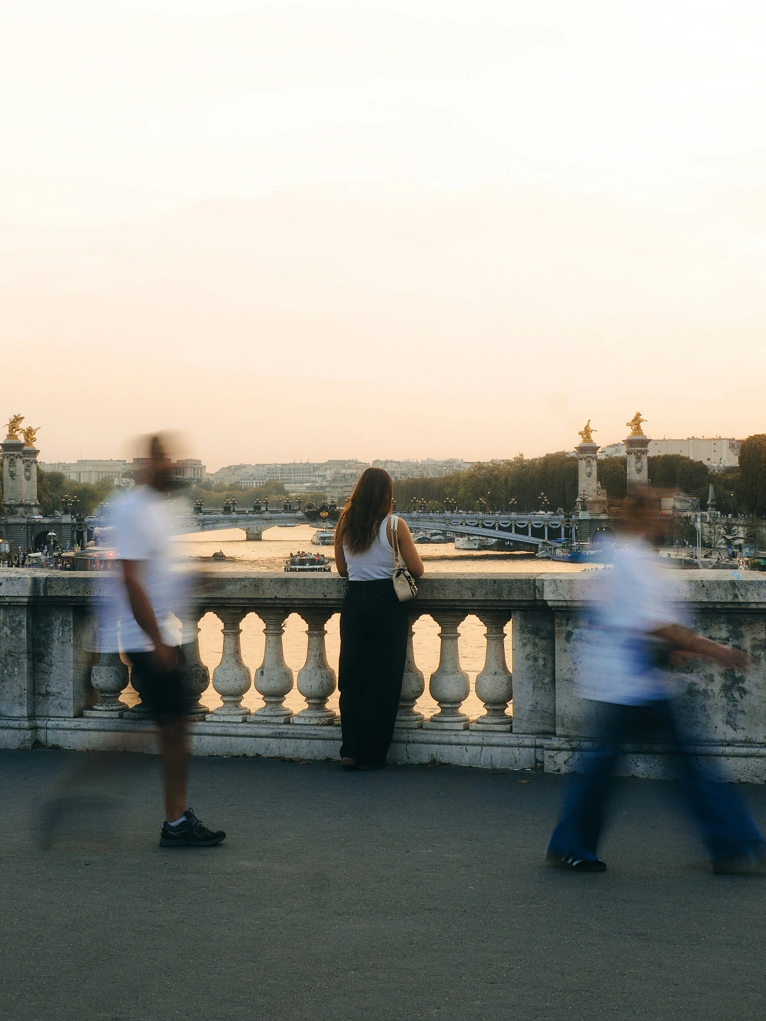 A woman with long hair stands by a stone railing overlooking a river at sunset, with a bridge and city buildings in the background. Blurred figures walk past in the foreground.