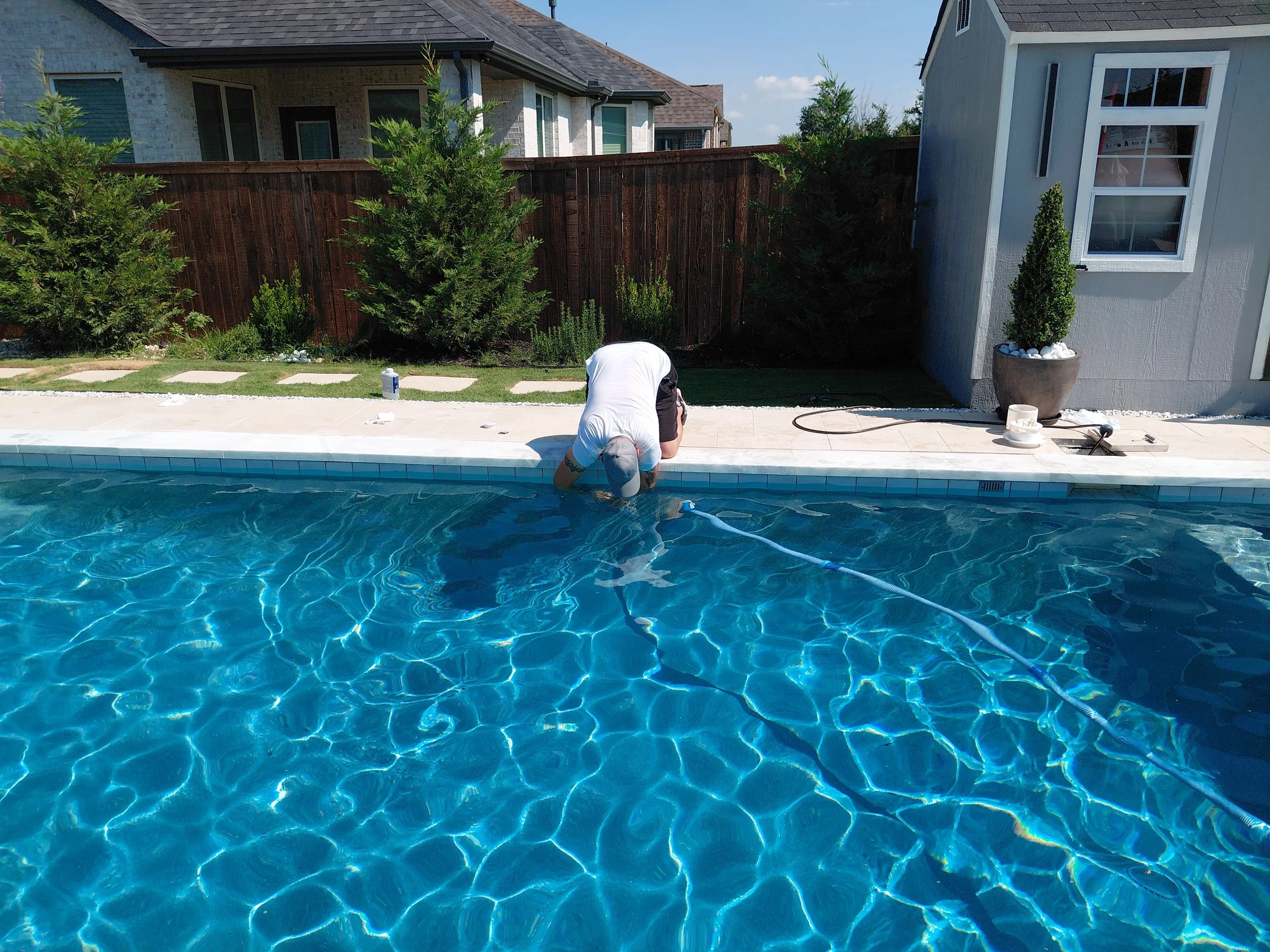 A person crouching by a swimming pool, wearing a white shirt and dark shorts, near a backyard with a wooden fence, green plants, and neighboring houses. The pool has blue water and clear reflections.
