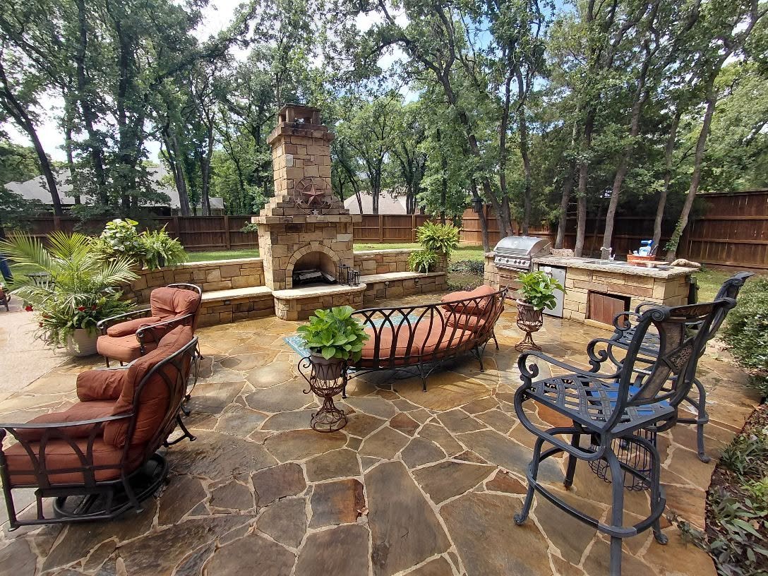 Backyard patio with stone flooring, outdoor fireplace, built-in grill, metal and cushioned seating, and lush trees in the background.
