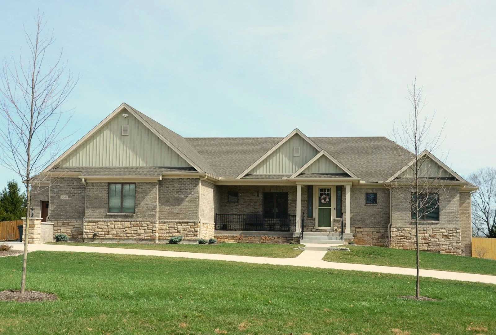 A single-family home with a mix of brick and stone exterior, small front porch, and a green front door with a wreath. Two leafless trees and a green lawn are in the foreground, with a clear sky above.