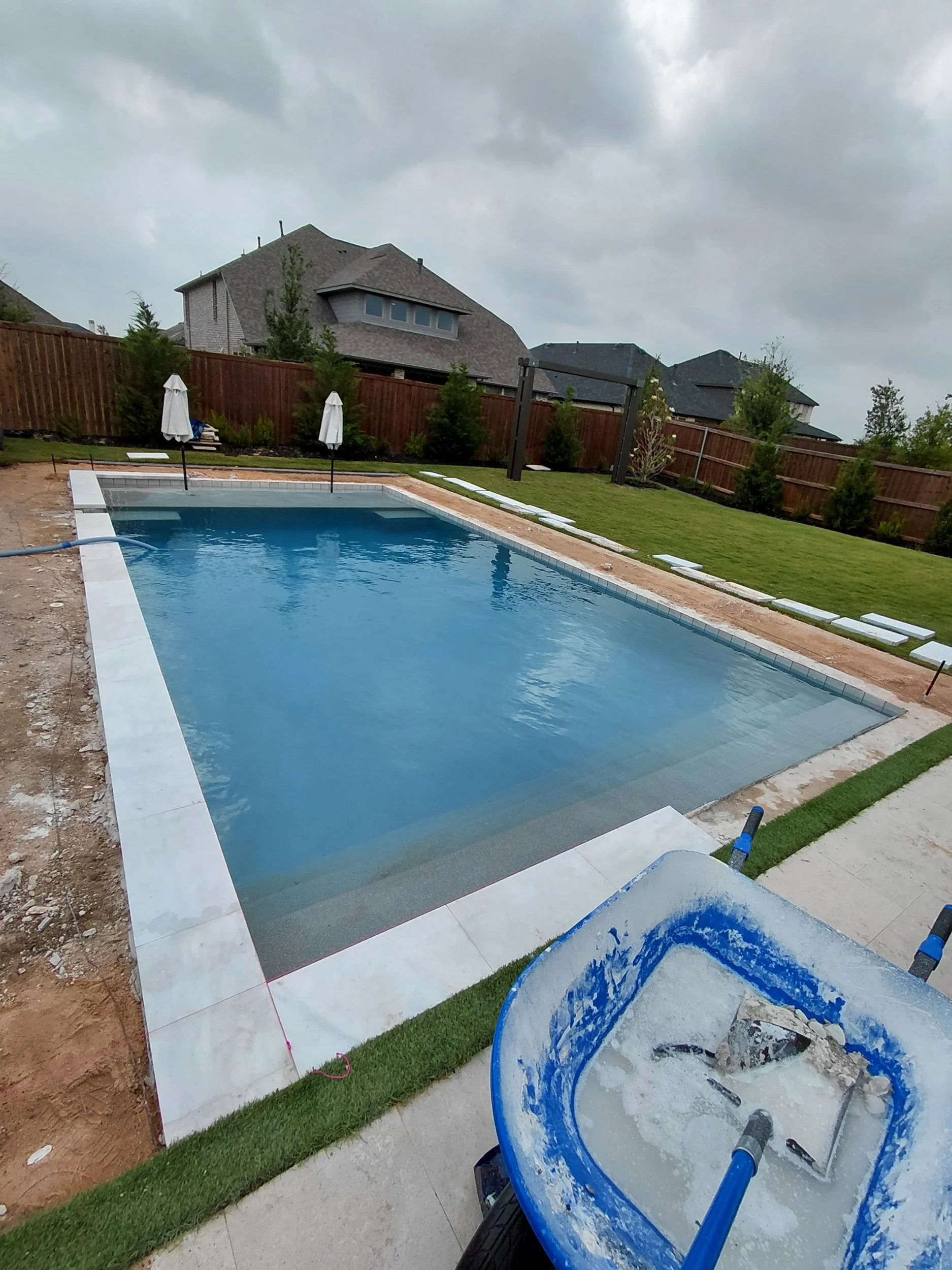 A backyard with a swimming pool under construction, with a blue pool filled with water, surrounded by a partially completed deck, green grass, a lawn, a wooden fence, and houses in the background under a cloudy sky.
