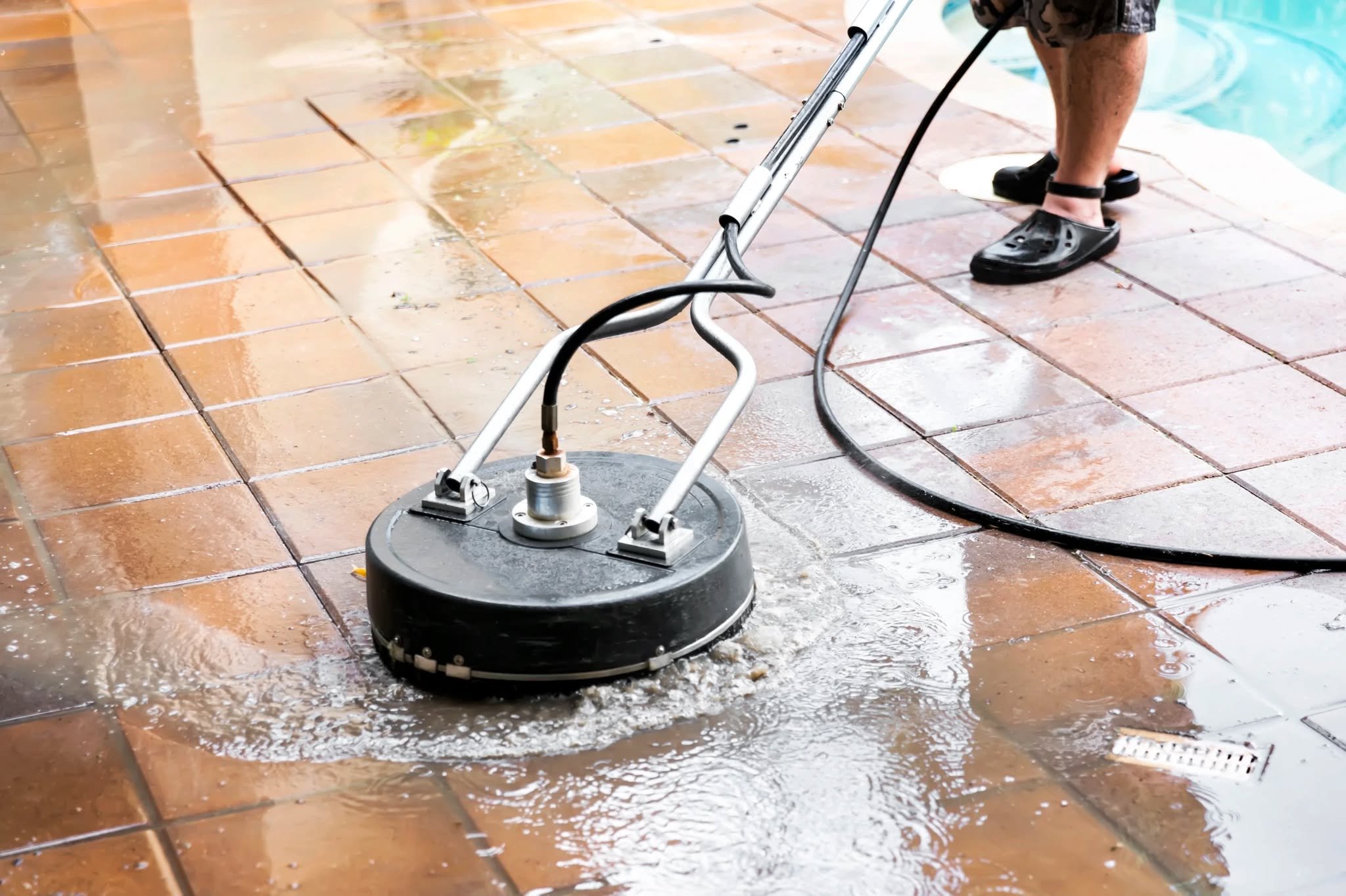 A person is using a wet floor sander on a tiled patio next to a swimming pool. The person is wearing black Crocs and is bent over while operating the machine, which is cleaning or polishing the tiles. Water and debris are visible on the tiles.
