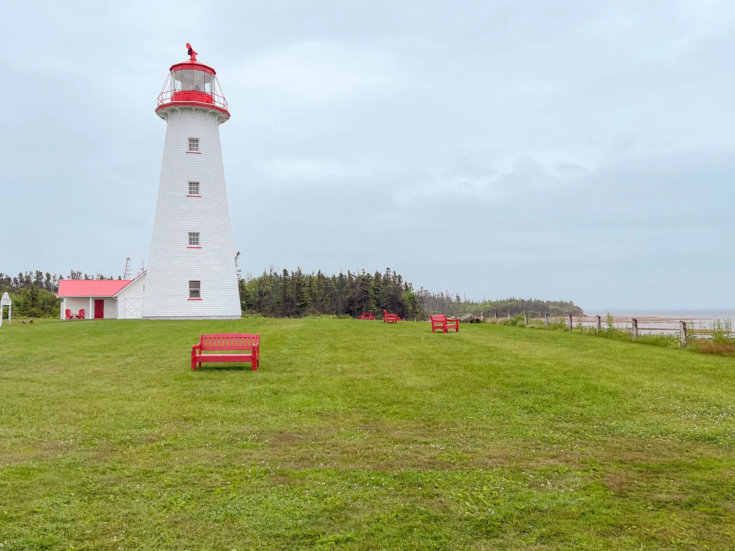 A white lighthouse with red accents on the top, windows, and benches surrounding it on a grassy area near the coast, with trees and a cloudy sky in the background.