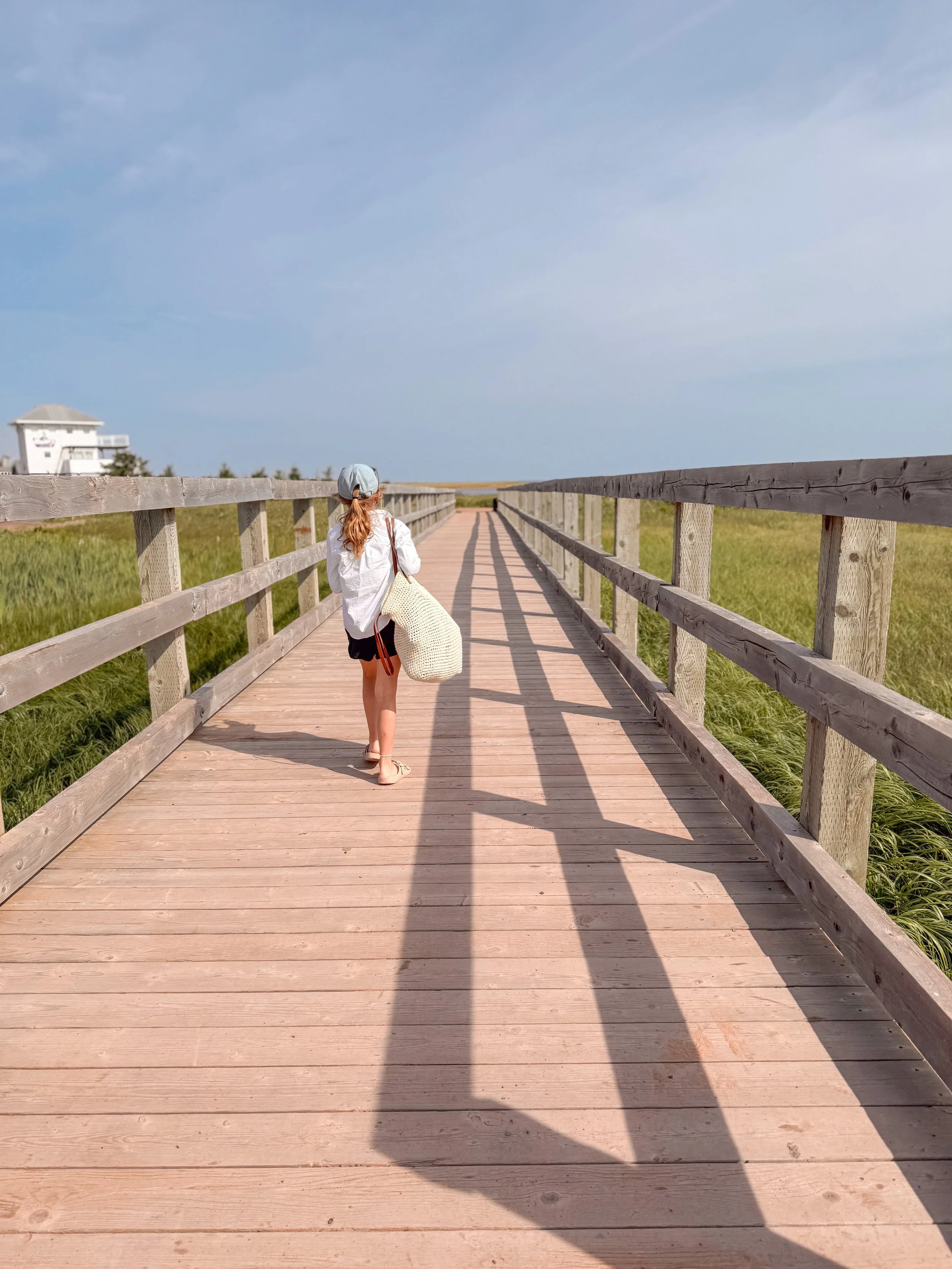 A woman with red hair wearing a white hat, white shirt, and black shorts walking on a wooden bridge with a railing in a grassy outdoor area on a sunny day.