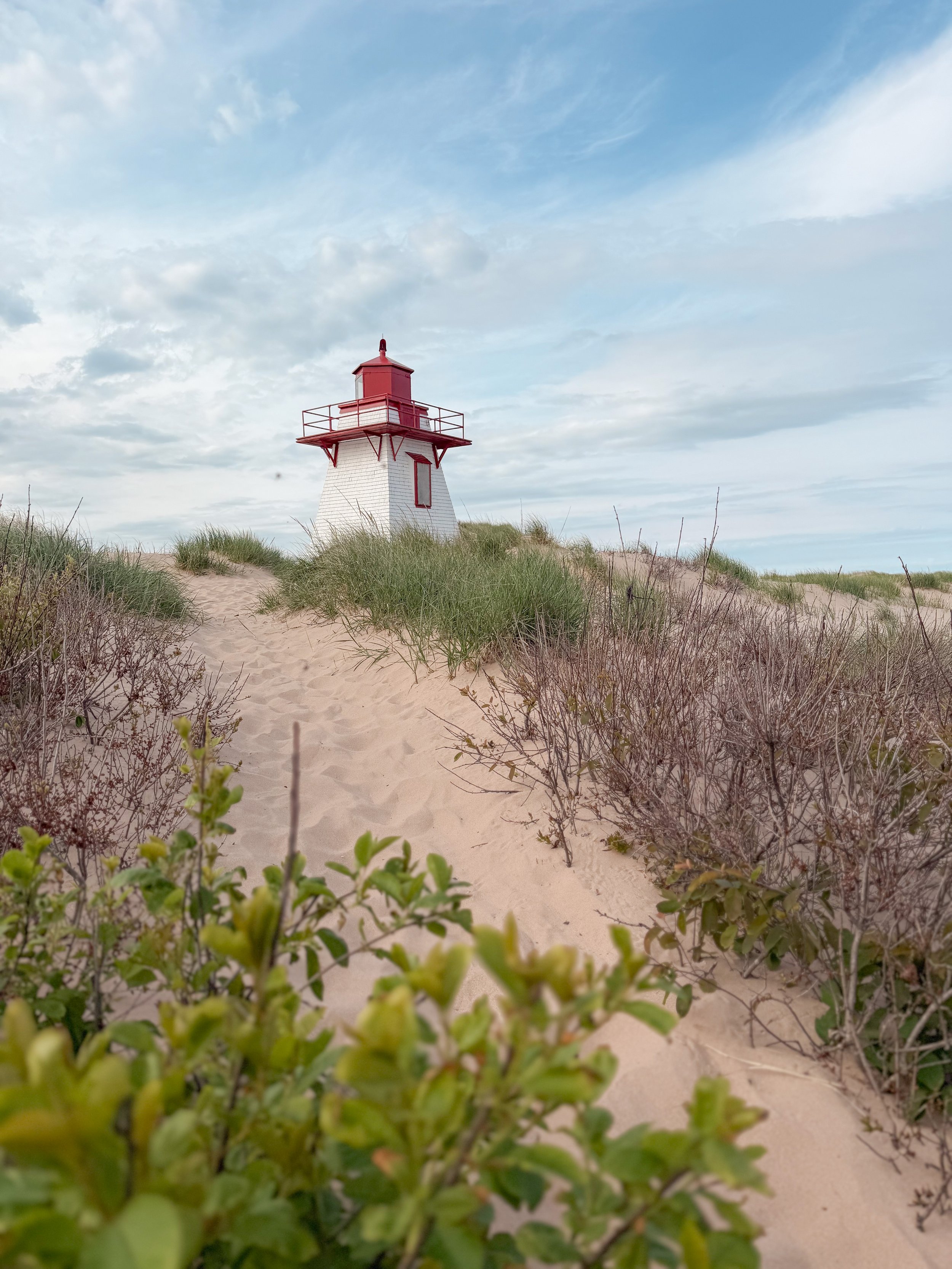 A white lighthouse with a red top and red railing, situated on sandy dunes covered with green grass and bushes under a partly cloudy sky.