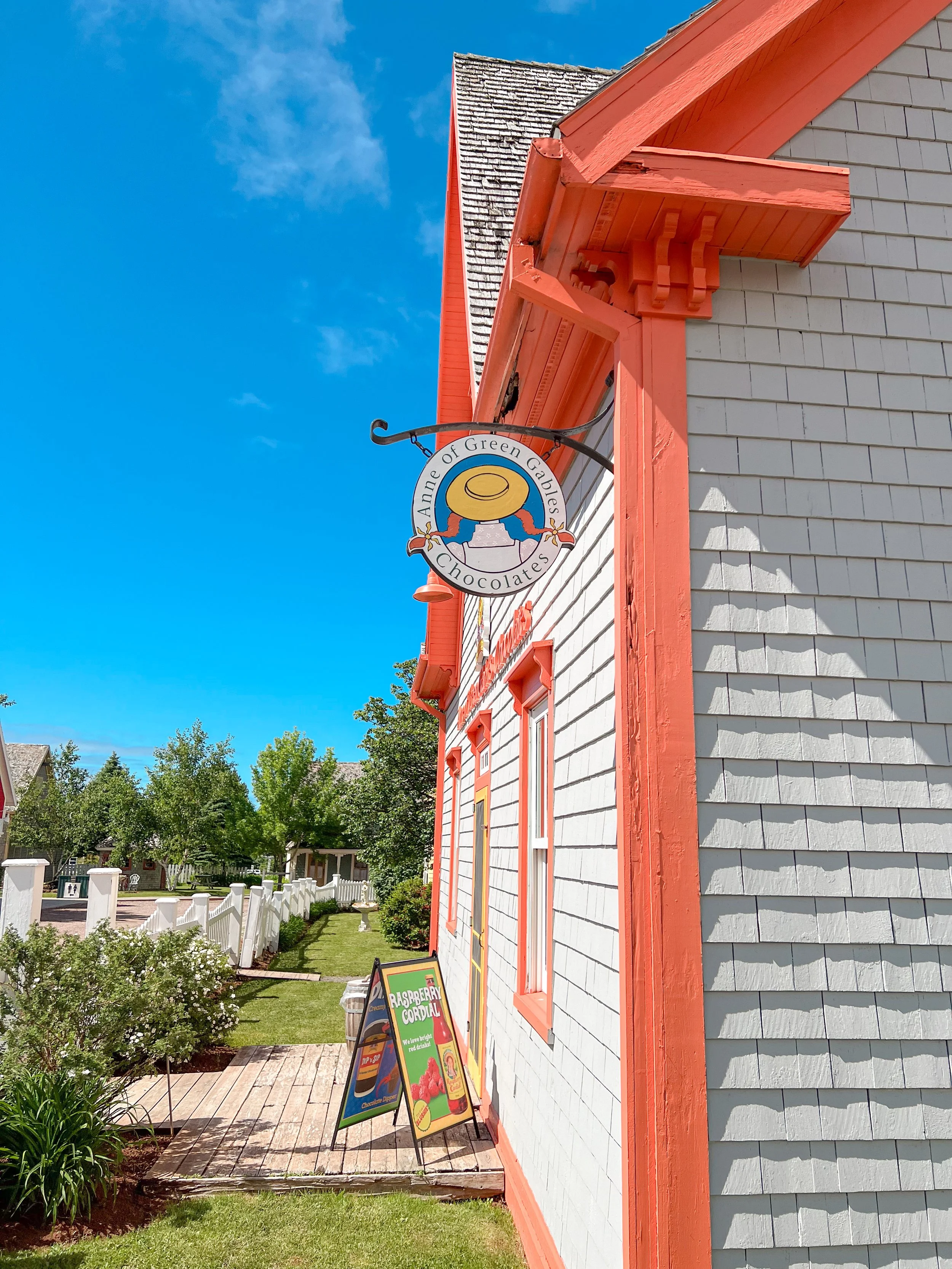 Close-up of a building’s exterior with gray siding and orange trim, a hanging sign for 'Anne of Green Gables Chocolates,' green trees and a fence in the background, and a signboard advertising raspberry cordial in front.