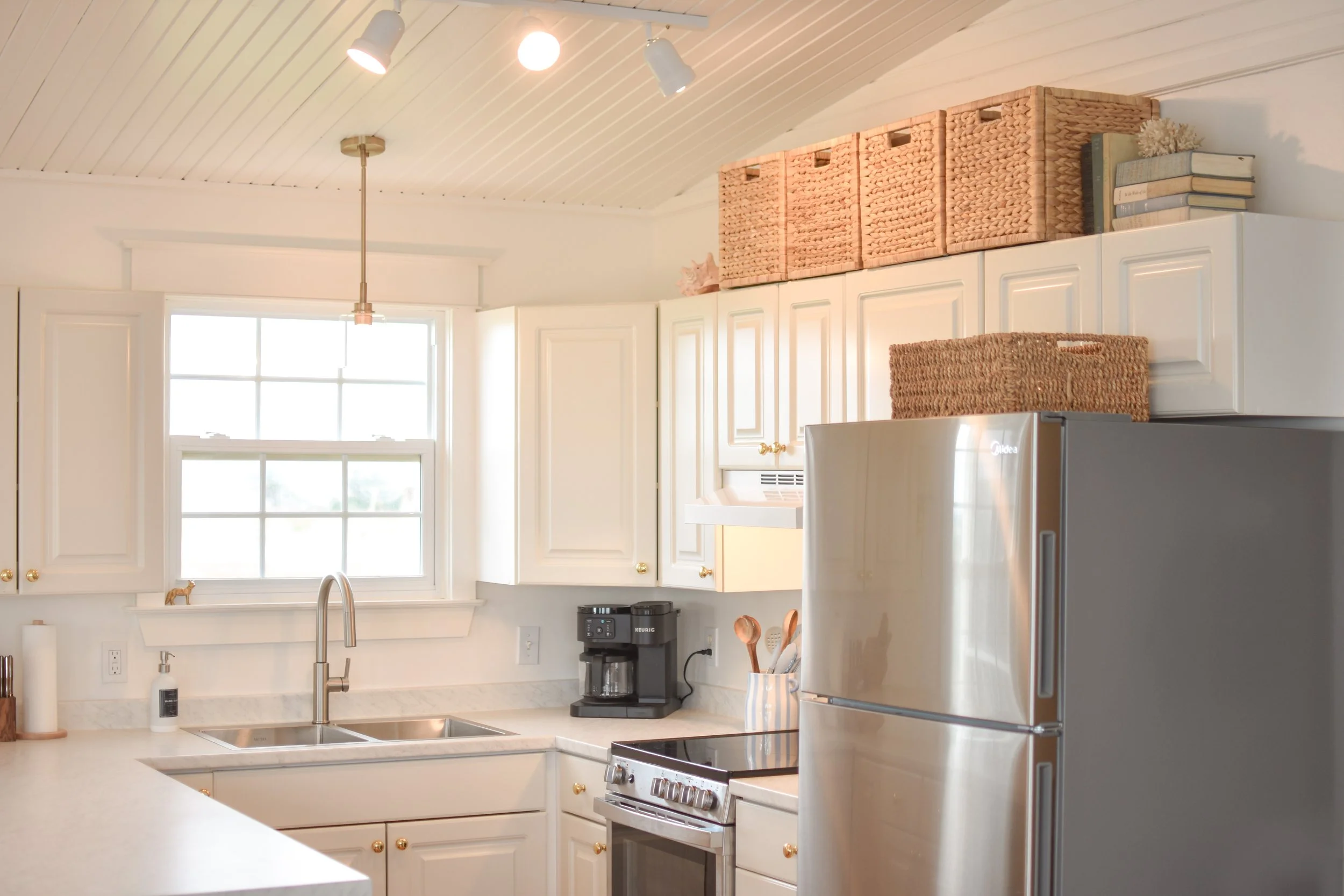 Kitchen with white cabinets, a window, a stainless steel refrigerator, a stovetop, and countertop with a coffee maker, utensils, and decor including woven baskets and books on top of the cabinets, and track lighting on the ceiling.