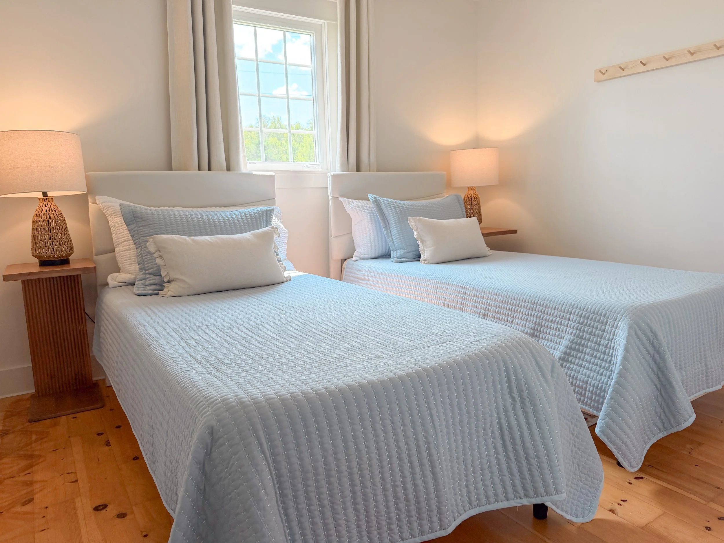 A bedroom with two beds, each with white bedspreads and pillows, wooden nightstand with wicker lamp, window with white curtains, and a wall hook rack.