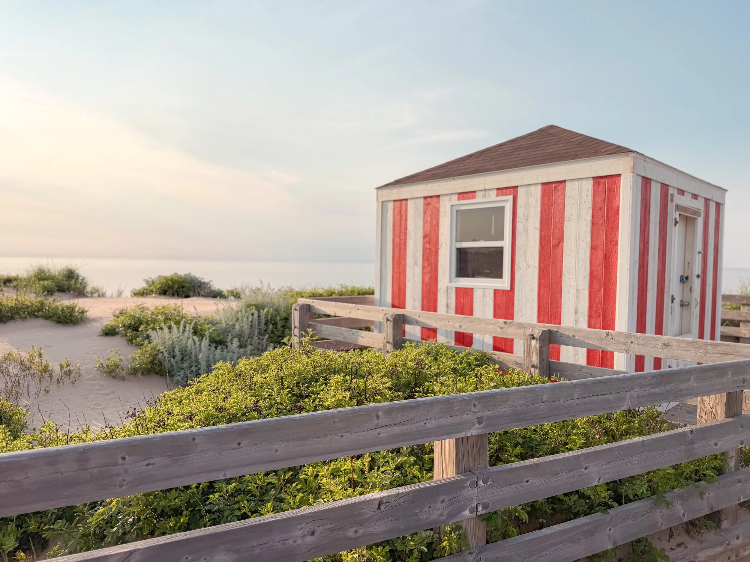 A small, red and white striped beach house on a sandy area with green bushes and a wooden fence in the foreground, under a clear sky.