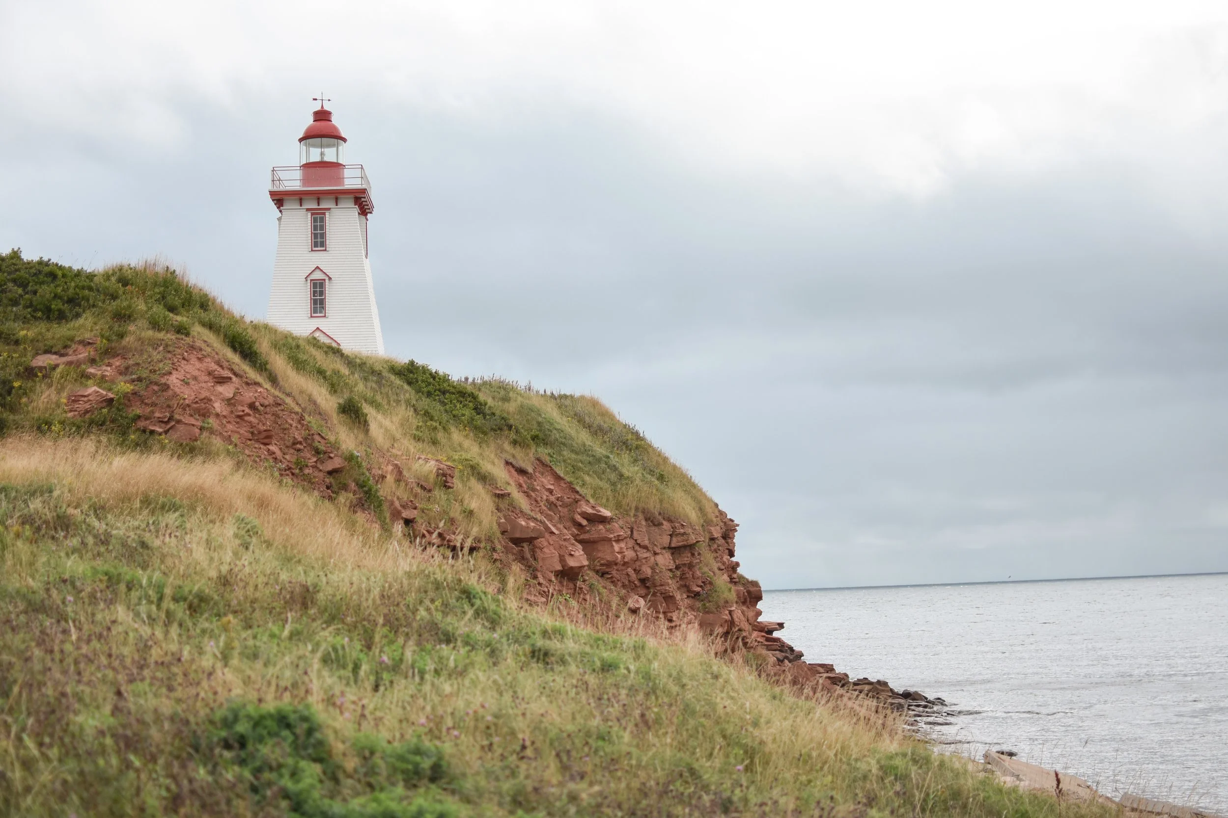A white lighthouse with a red top and railing, situated on a grassy, rocky hillside by the sea under an overcast sky.