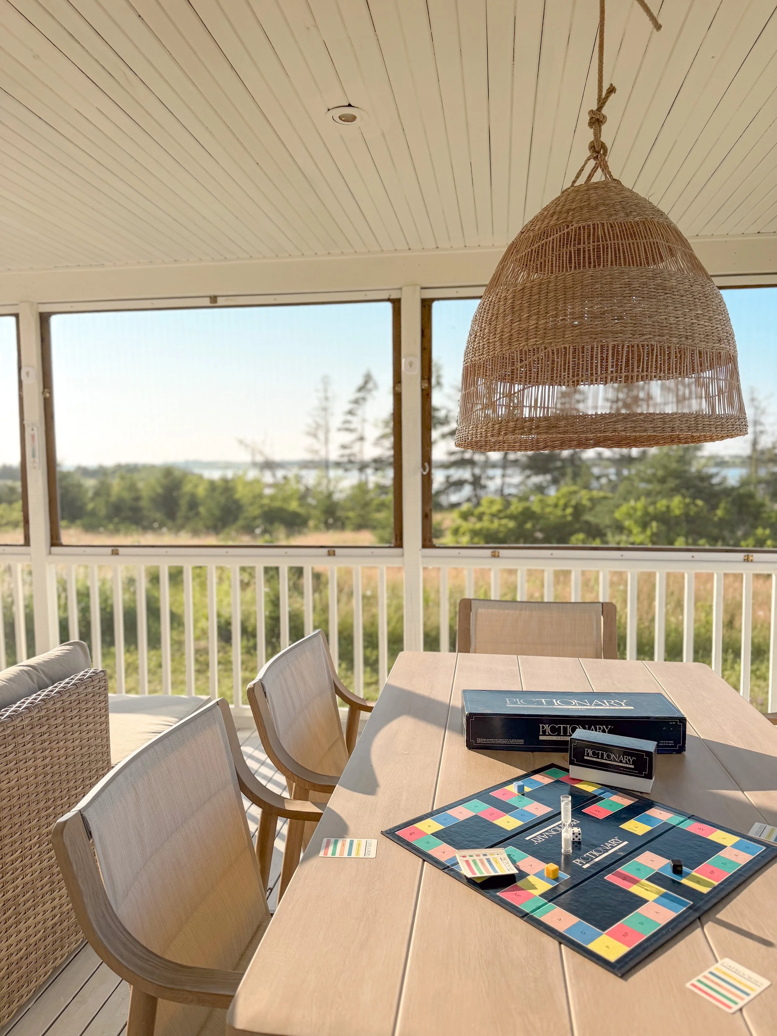 A living room with large windows, a wooden dining table with board games, and chairs. There is a woven lampshade hanging from the ceiling, and outside there are trees and a view of water in the distance.