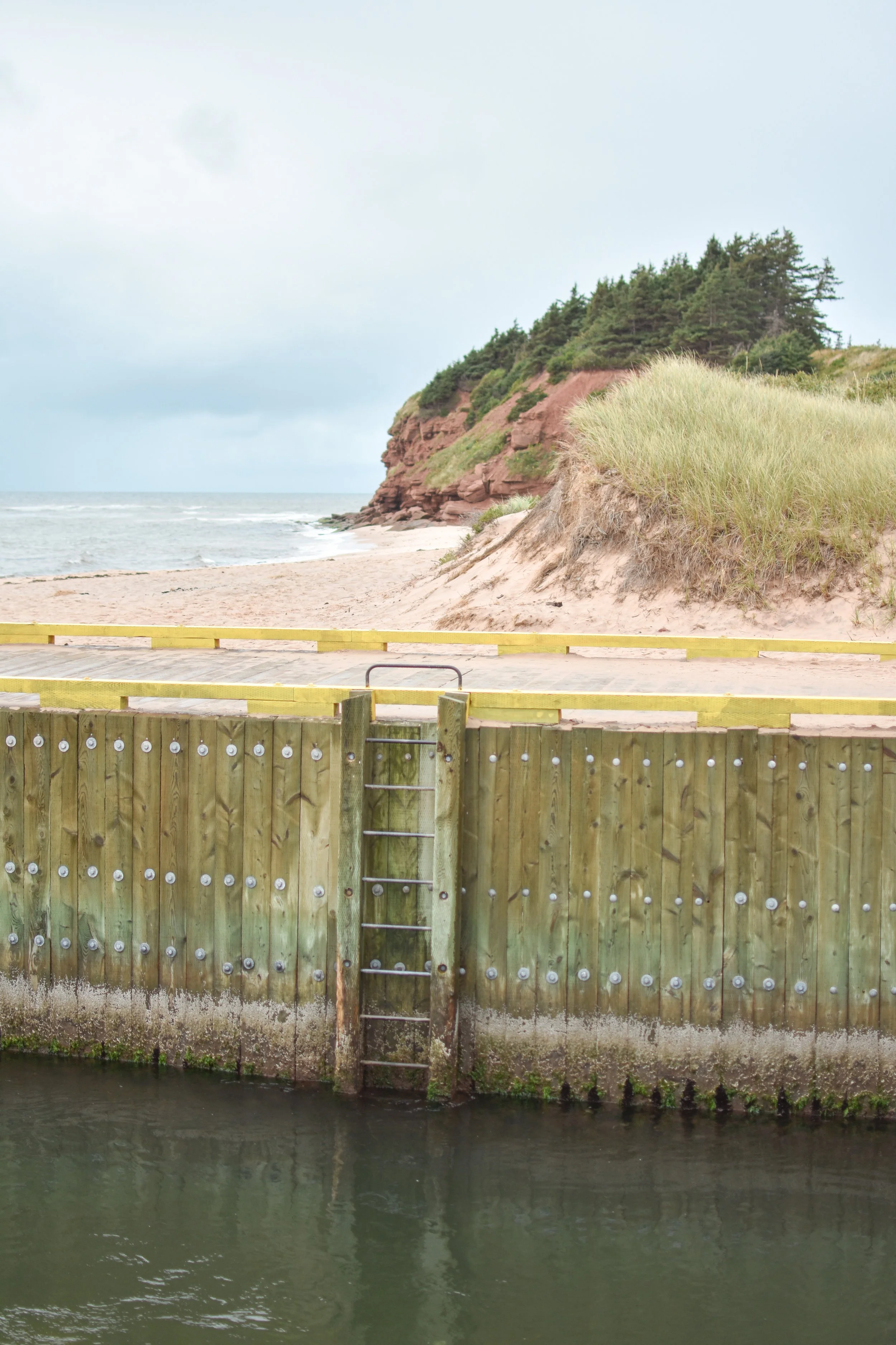 A view of a sandy beach with a rocky slope covered in trees, seen from behind a wooden fence along a canal or river. The water is calm with a cloudy sky overhead.