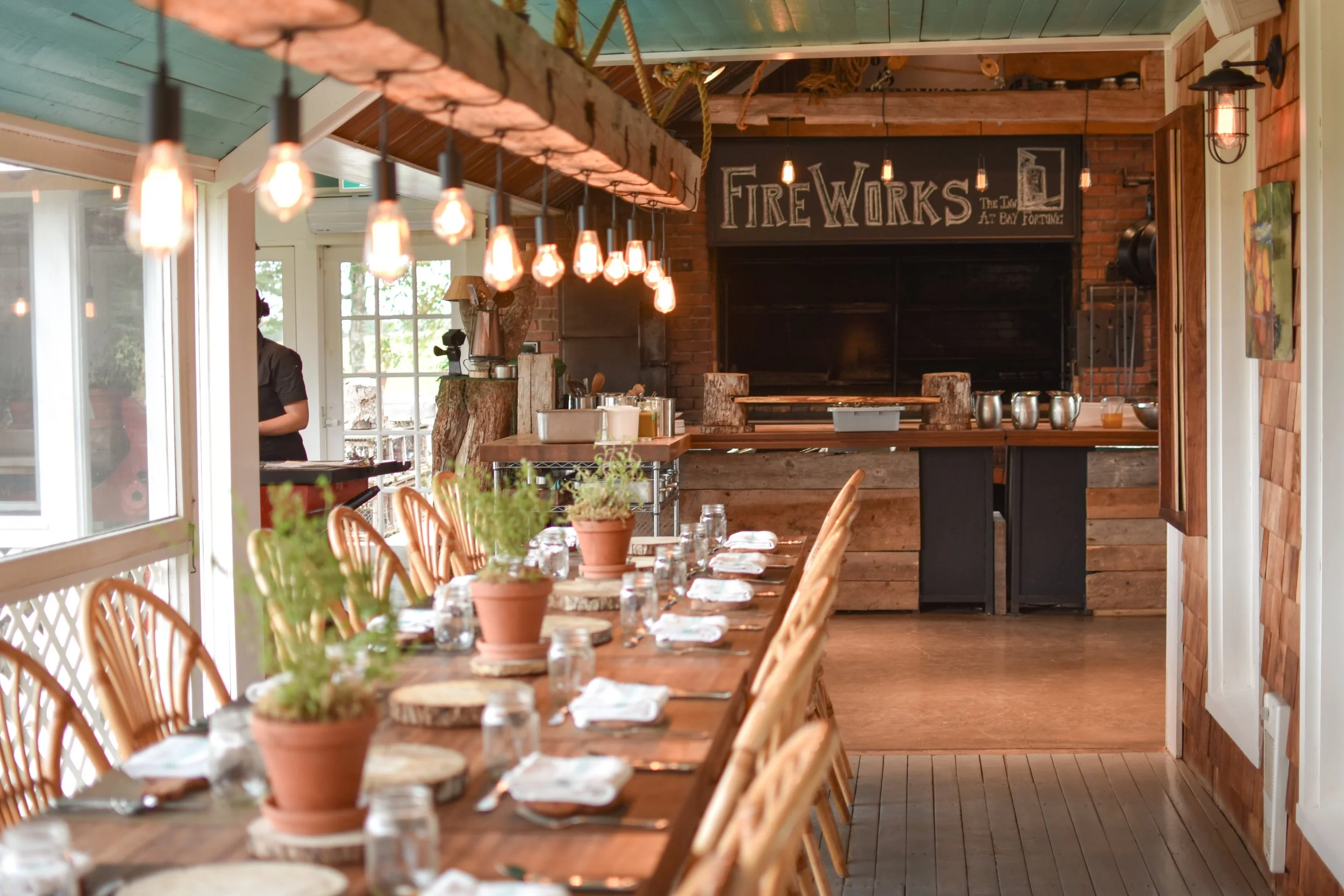 A rustic dining area with a long wooden table set for a meal, adorned with potted plants, glasses, and napkins. The room features large windows, wooden walls, and decorative hanging light bulbs. There is a sign that says 'Fireworks' in the background and a brick fireplace.