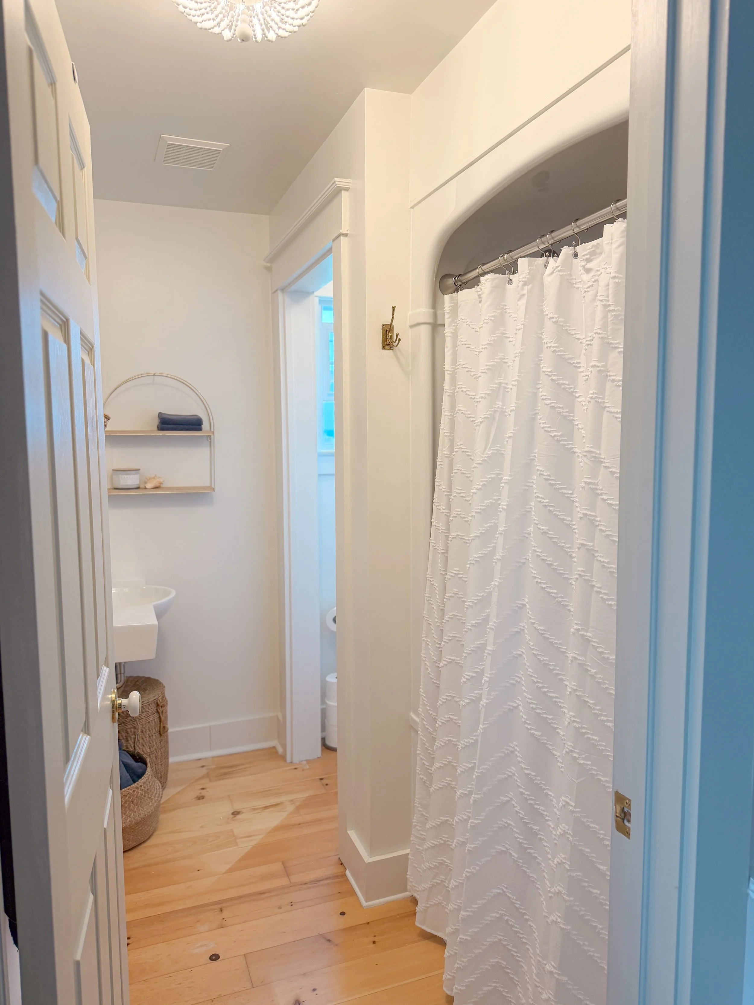 A small bathroom with a white patterned shower curtain, a niche with towels and shells, a small area for a toilet, and a hardwood floor.