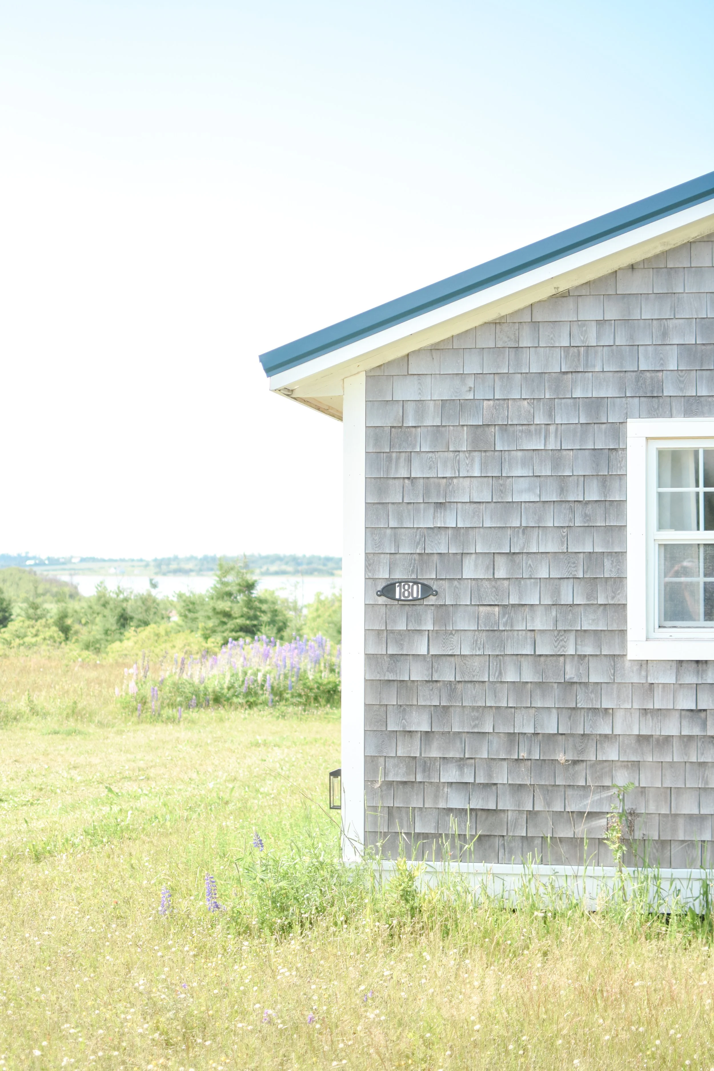 A house with gray wooden siding, a white window, and a black house number plaque reading 180, situated in a field of grass and purple flowers, with green trees and water in the distance under a clear blue sky.
