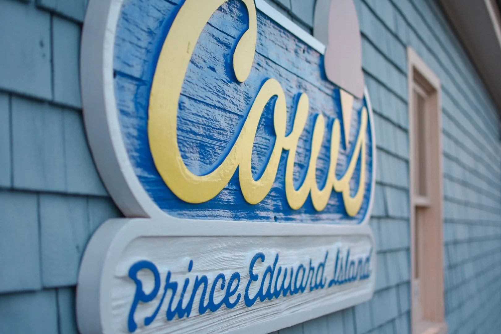 Close-up of a sign on a house with blue shingles, showing the name "Coves" in yellow script and the words "Prince Edward Island" in blue below it. The house has a window nearby.