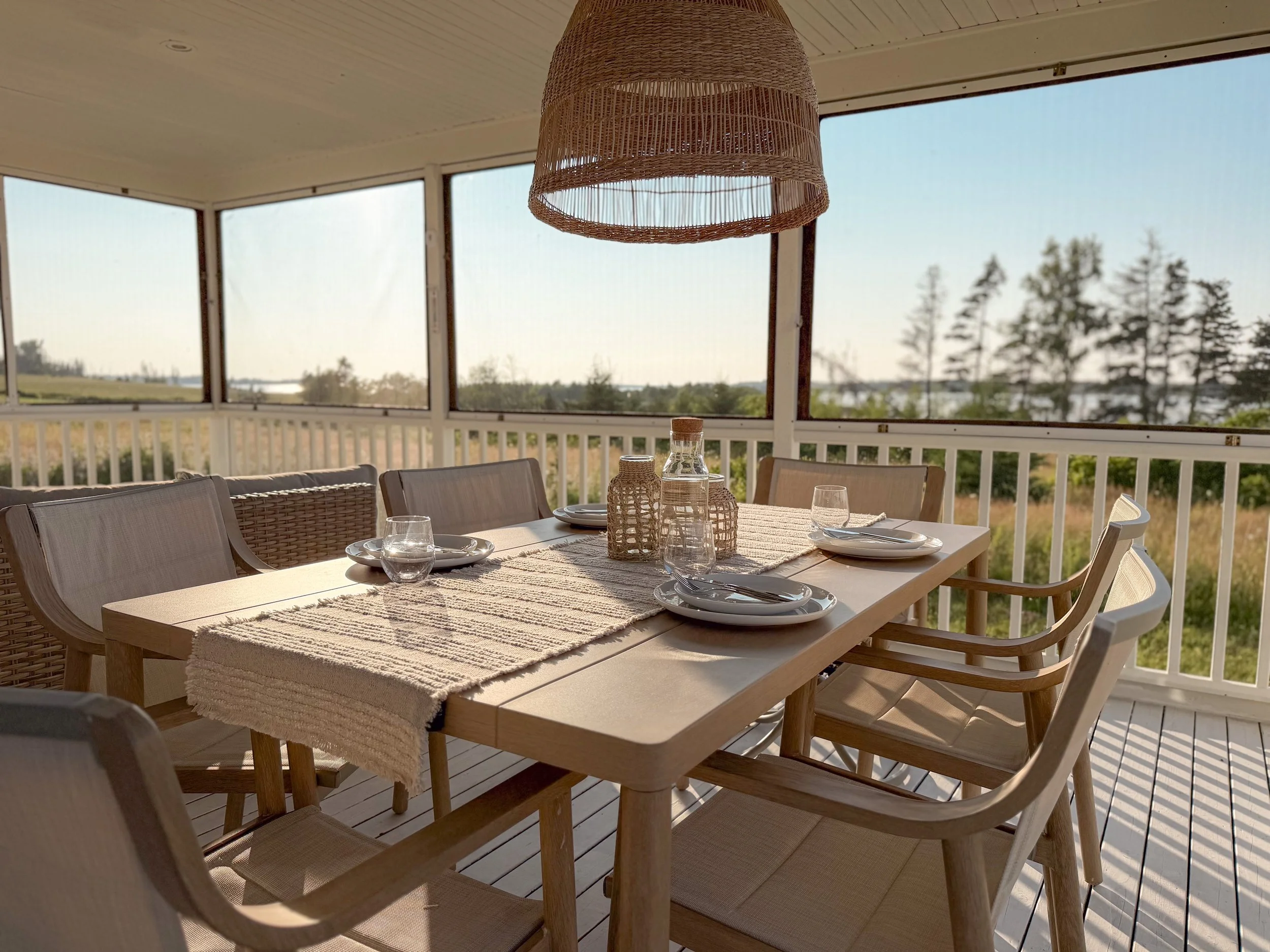 A dining table set with plates, glasses, and woven centerpieces, located on a covered porch with large windows overlooking a landscape with trees and water in the distance.