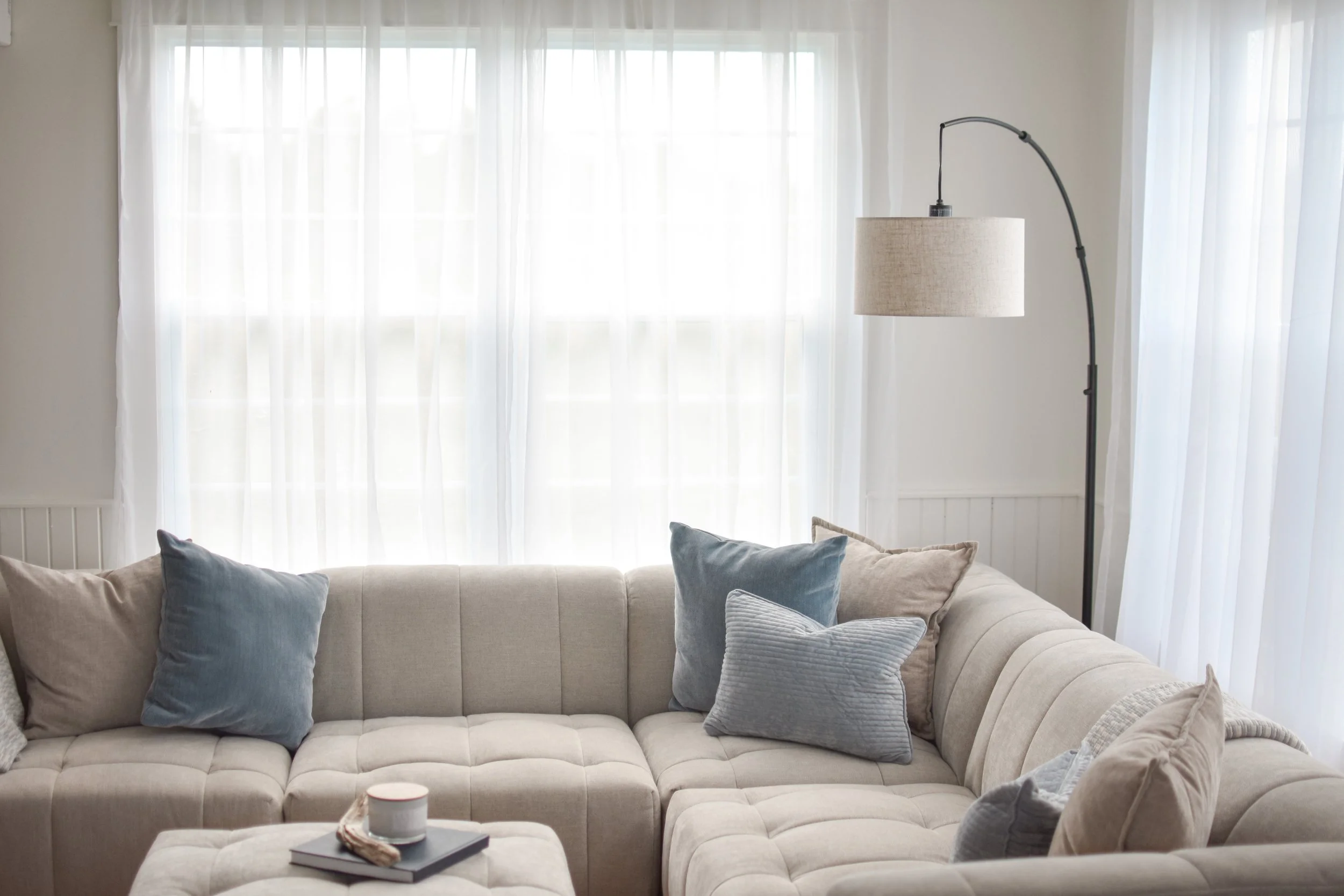 A cozy living room corner with a beige tufted sectional sofa adorned with blue and grey pillows. Behind it, large white sheer curtains allow natural light into the room. A floor lamp with a beige lampshade stands next to the sofa, and a coffee table with a cup, book, and remote is in front of the sofa.