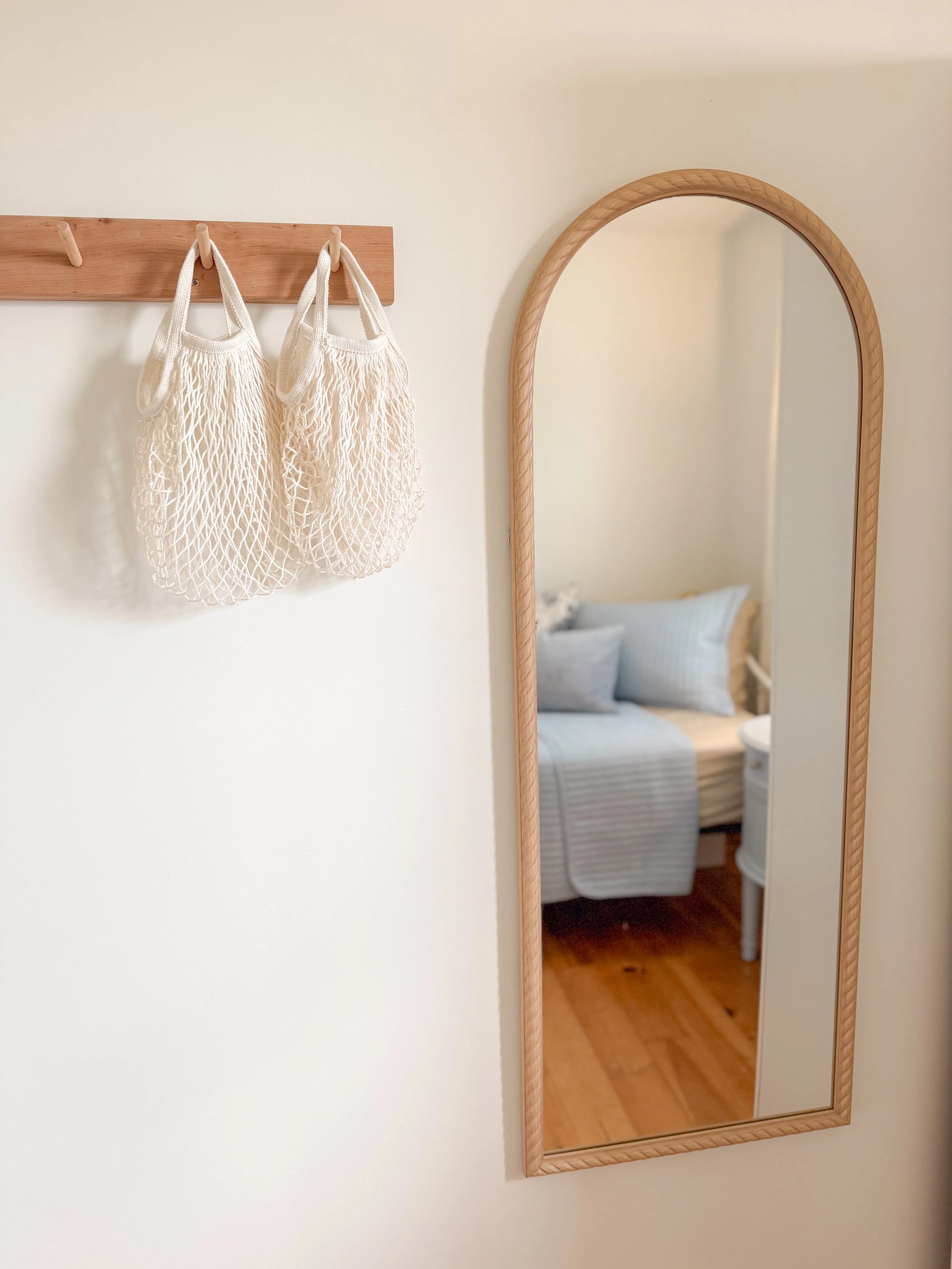 A wooden coat rack with two beige mesh shopping bags hanging on it, and a tall wooden framed mirror reflecting a room with a bed, pillows, and a small side table.