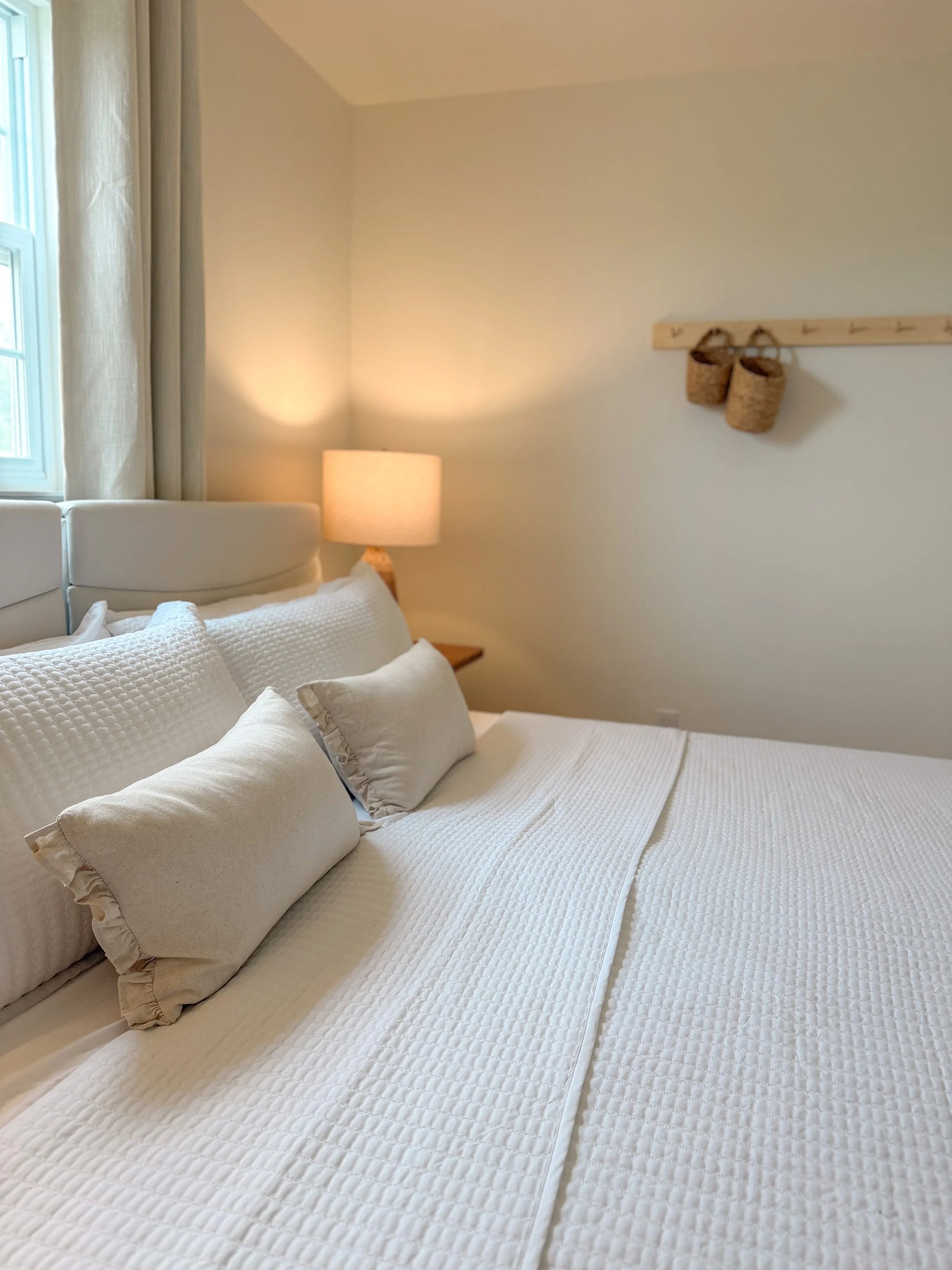 A cozy bedroom corner with a bed, beige pillows, a window with curtains, a side table with a lamp, and a wall hook with two storage baskets.