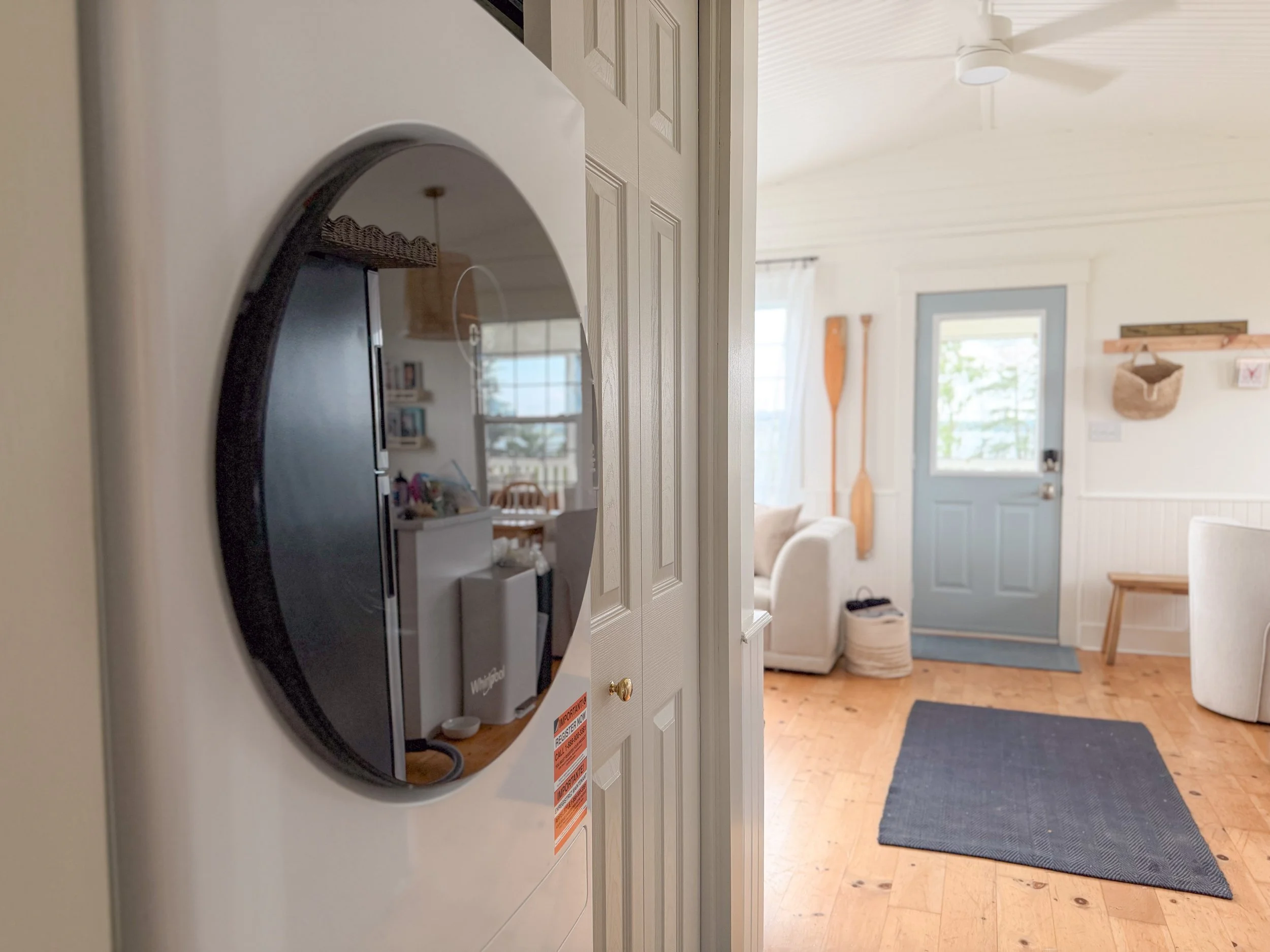A laundry room with a front-loading washer on the left side, with its door open revealing the interior. The room has a door with a window, a blue rug on a wood floor, an armchair, a small basket, and wall decorations including a paddle and a hanging bag.