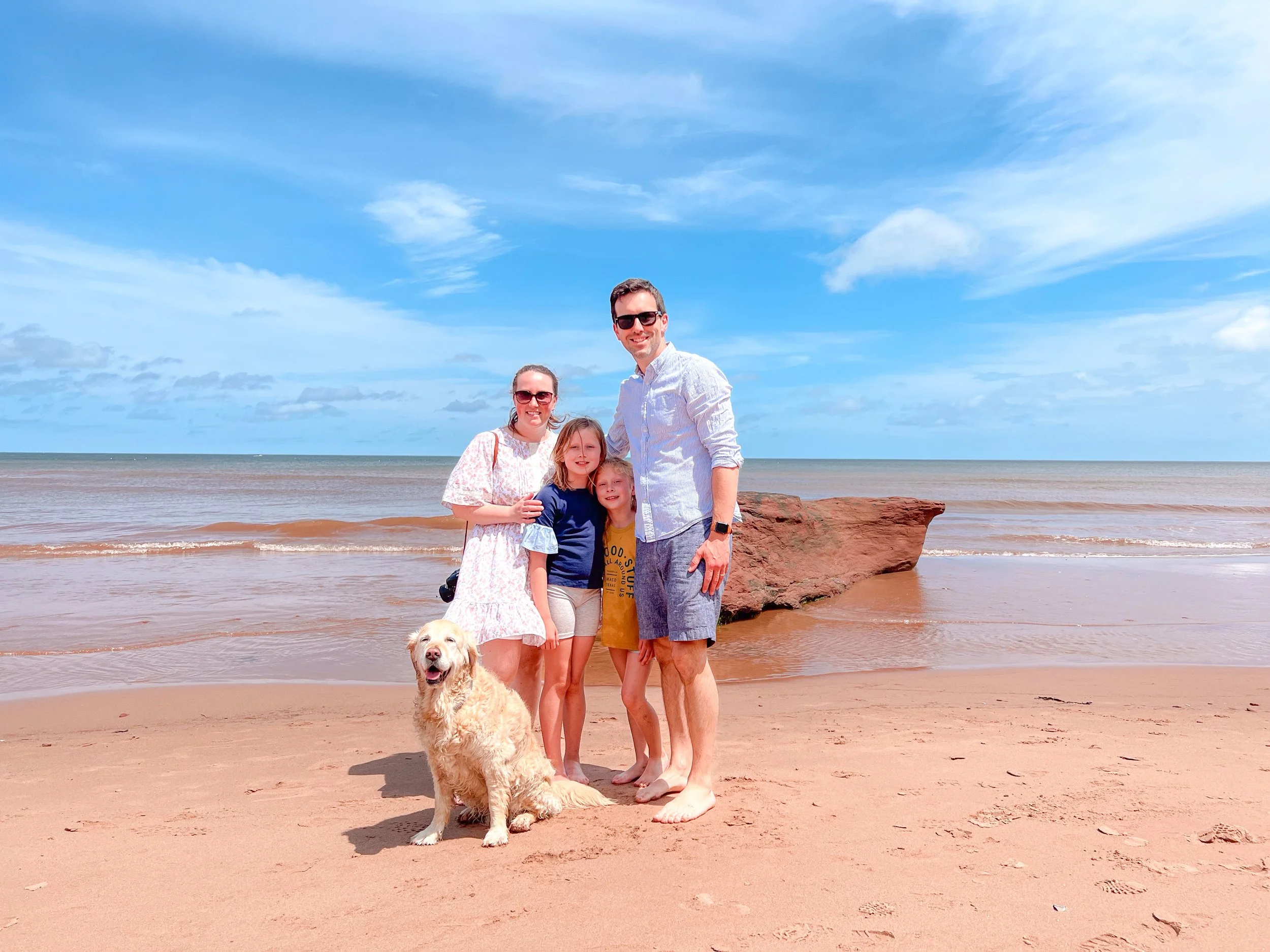 A family of four with two young children and a dog on a sandy beach with ocean waves in the background under a partly cloudy blue sky.