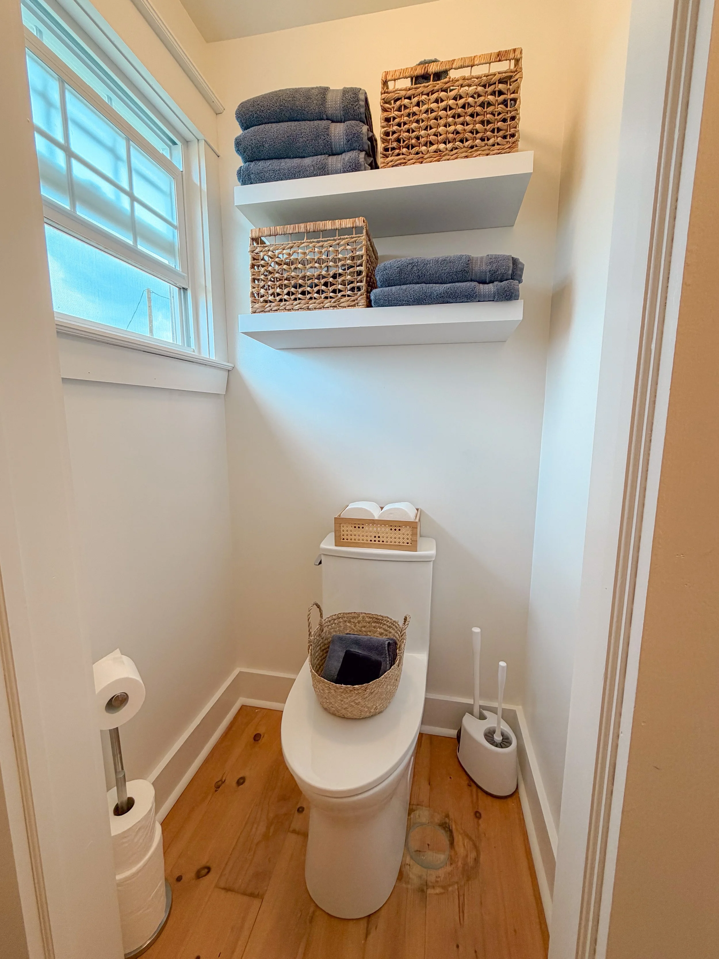 Small bathroom with a toilet, wicker basket on tank, window with blinds, two white shelves above the toilet with towels and baskets, toilet paper and cleaning tools nearby, wooden floor.